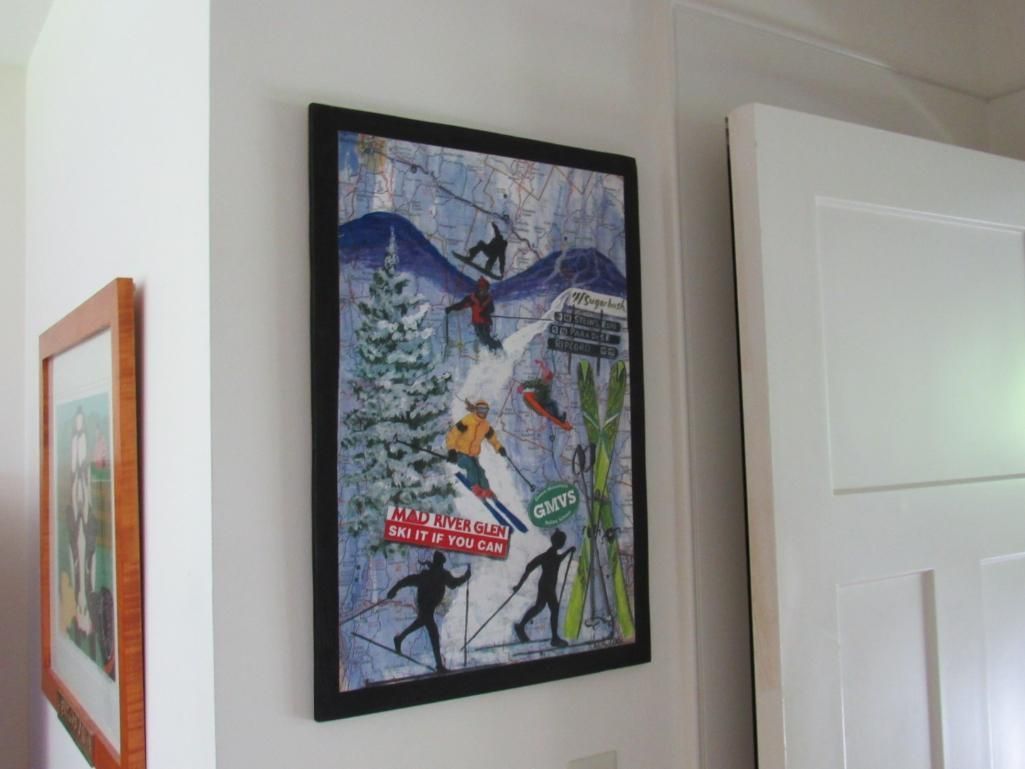 Skiing poster on wall with skiers and mountain scene. Black frame, door and smaller framed artwork visible.