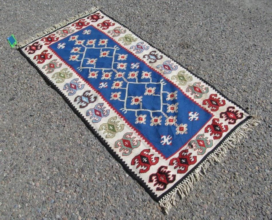 Rectangular rug on pavement with blue, red, white, and green geometric patterns and fringe.