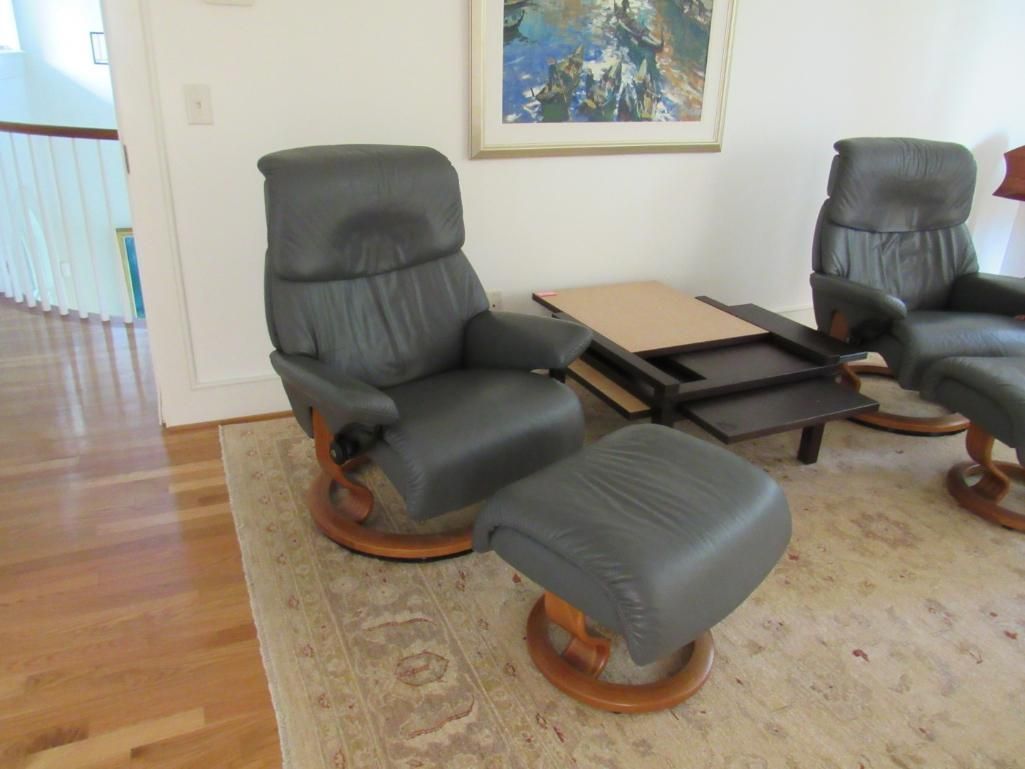 Two gray leather recliners and ottoman on a patterned rug in front of a painting.