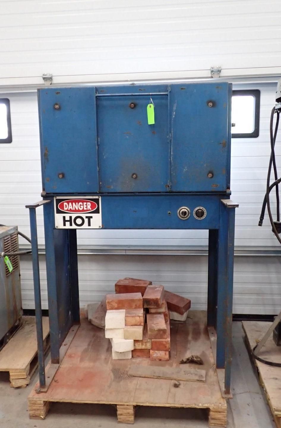Blue industrial oven on a metal stand with a warning sign. Bricks are on a wood pallet underneath.