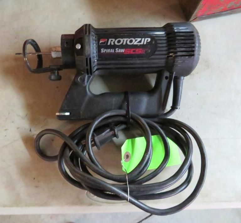 Black Rotozip spiral saw with cord, resting on a surface. A green tag is attached to the cord.
