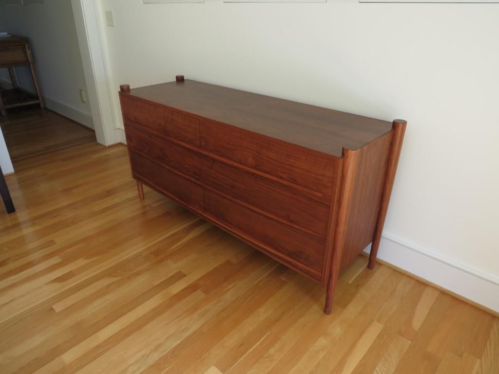 Brown wooden chest of drawers on a wooden floor, set against a white wall.