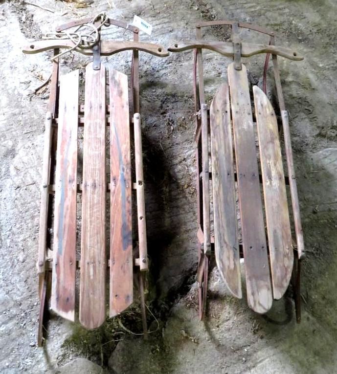 Two vintage wooden sleds with metal runners and steering mechanisms, leaning against a rough surface.