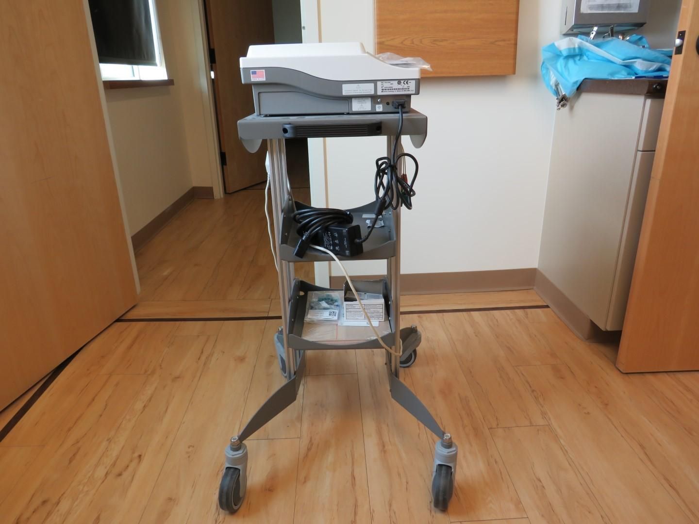 Medical equipment cart on wheels, holding a machine with cords and supplies, in a room.