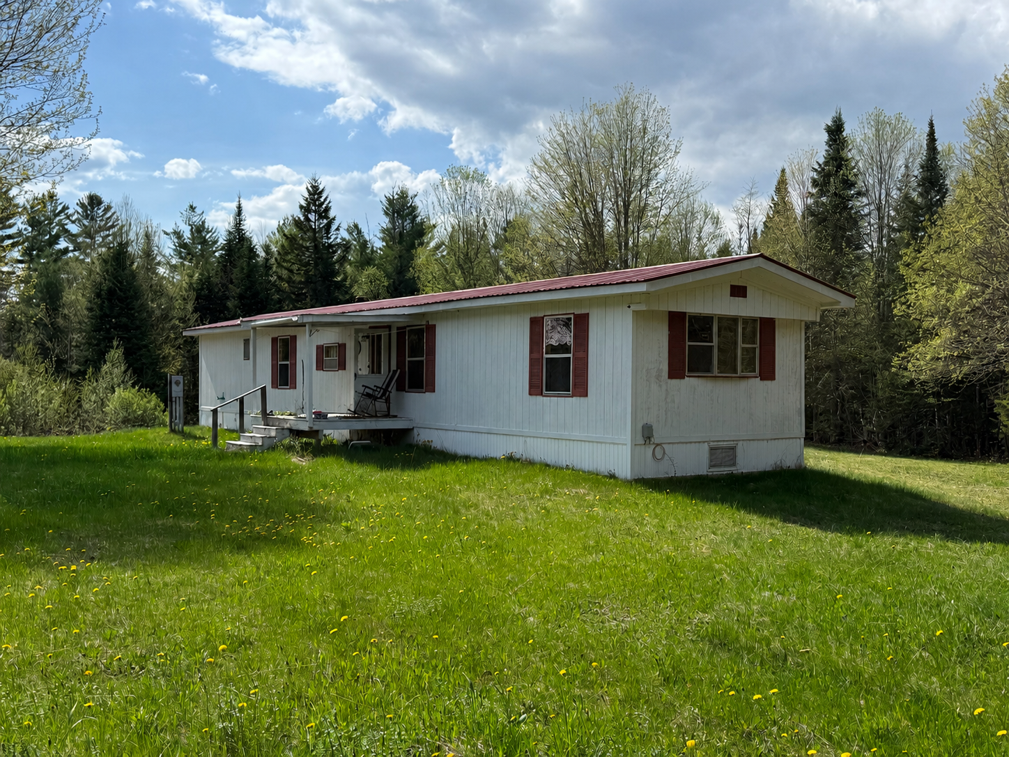 White single-story house with red roof on a grassy lawn, surrounded by trees under a blue sky