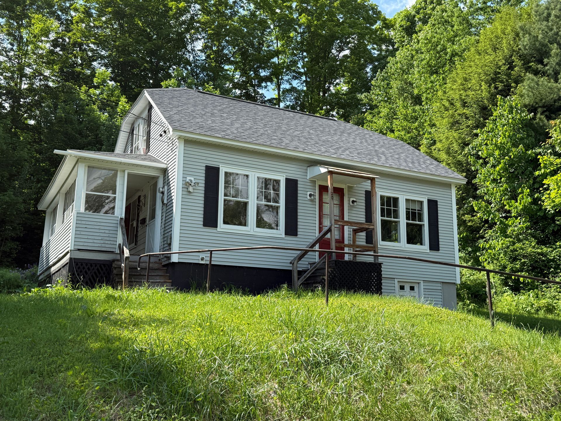 Small white house with a porch and red ladder on a grassy, wooded hillside