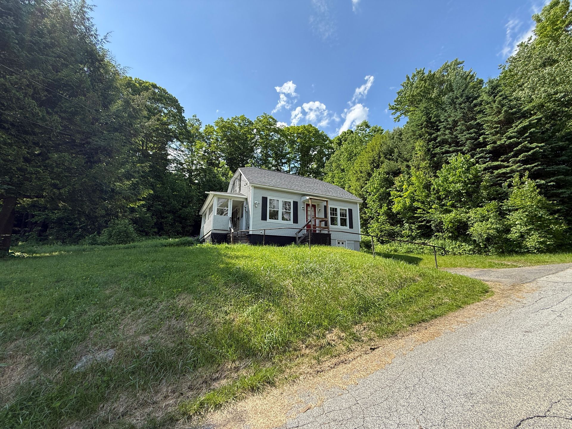 Small white house with porch beside a gravel road, surrounded by green trees and grass under a blue sky