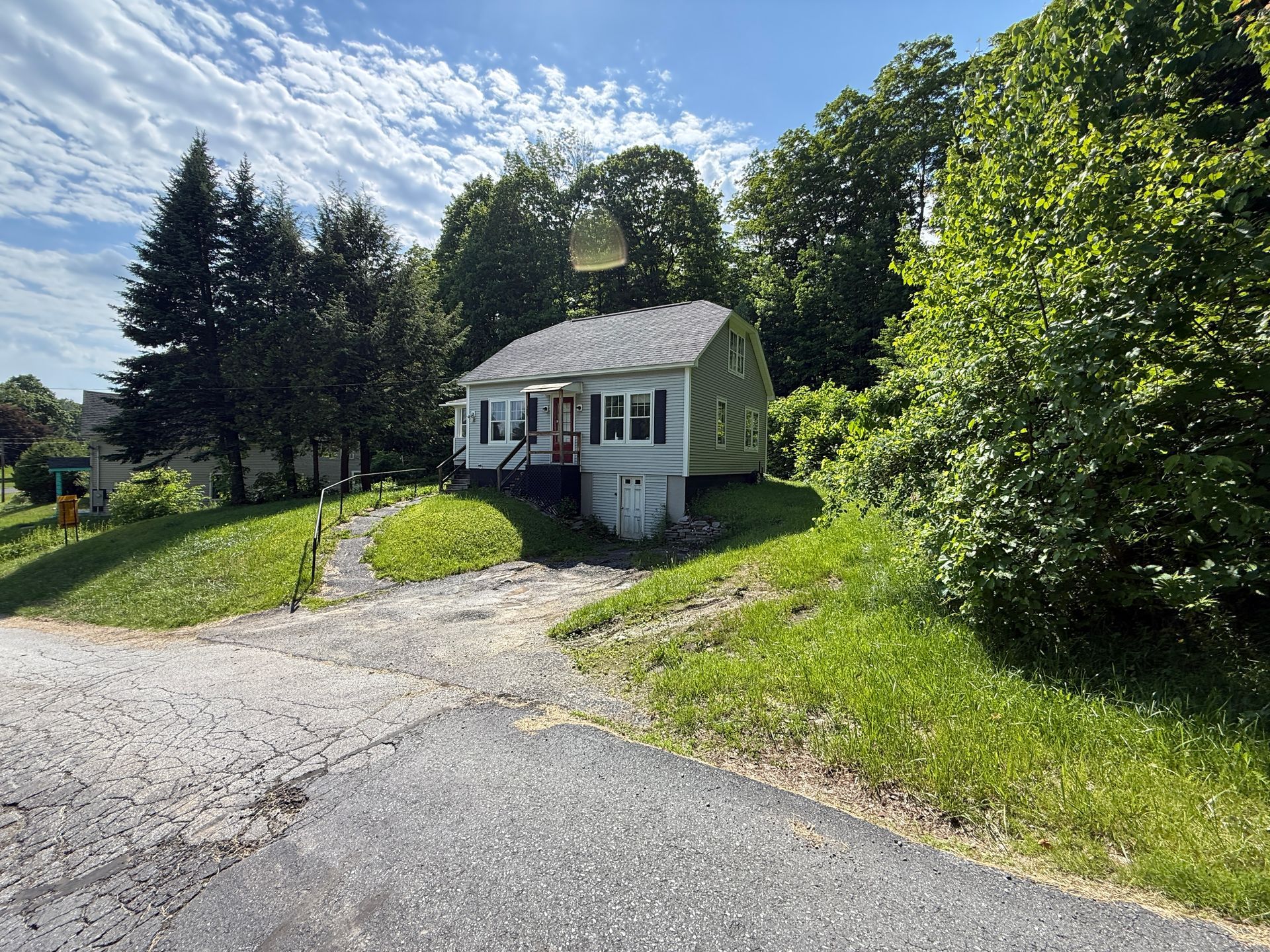 Small white house at the end of a gravel driveway, surrounded by green trees and lawn under a bright blue sky.