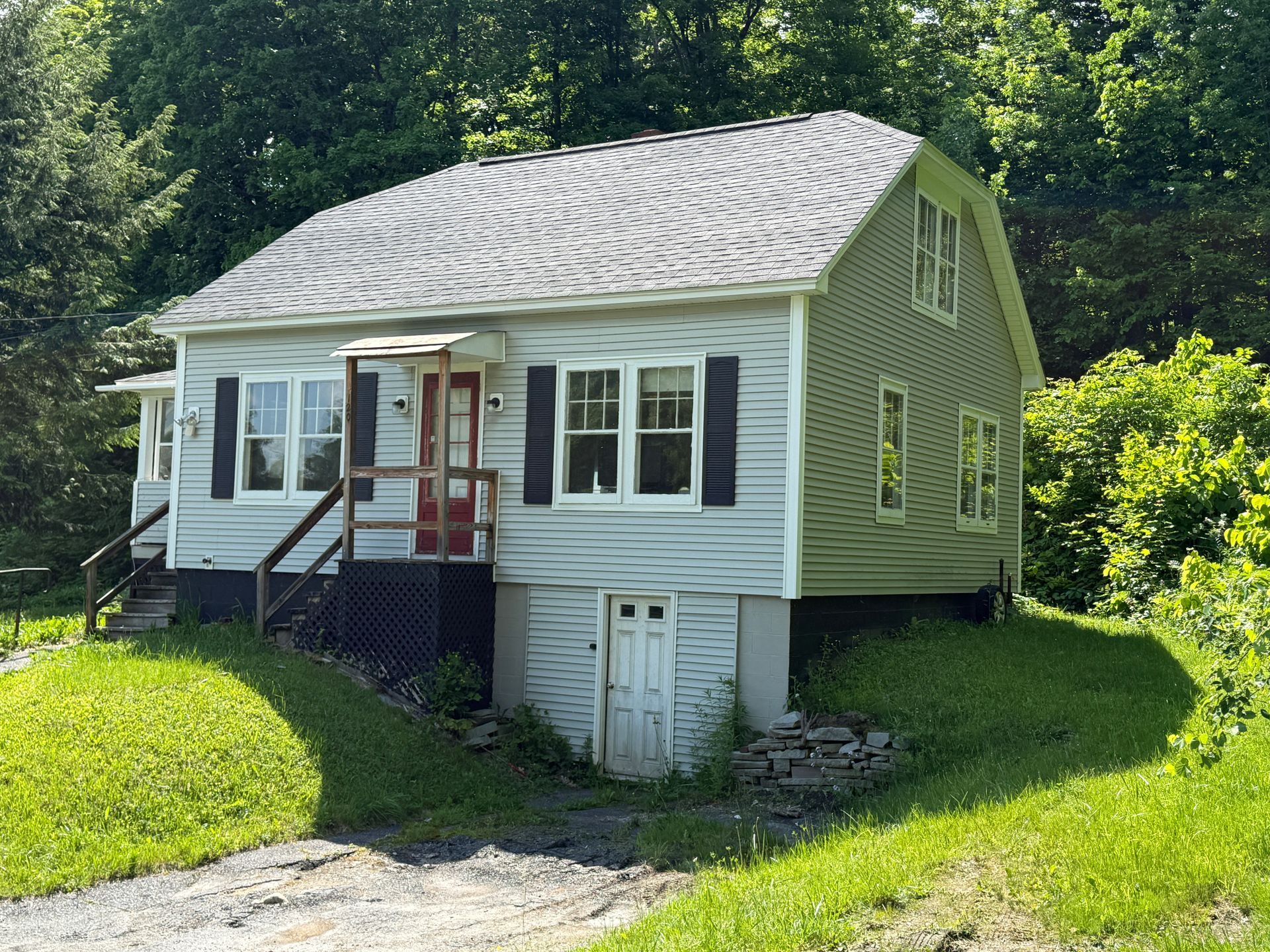 Small green house with white trim and a front porch on a grassy hillside by trees