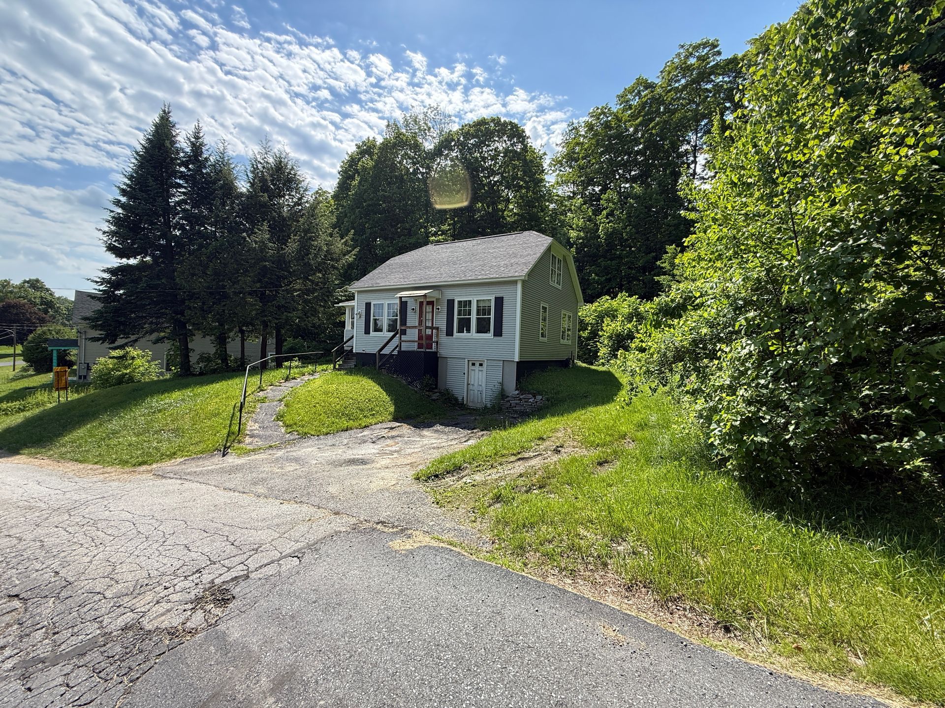 Small white house with a porch beside a driveway, surrounded by green trees and grass on a sunny day
