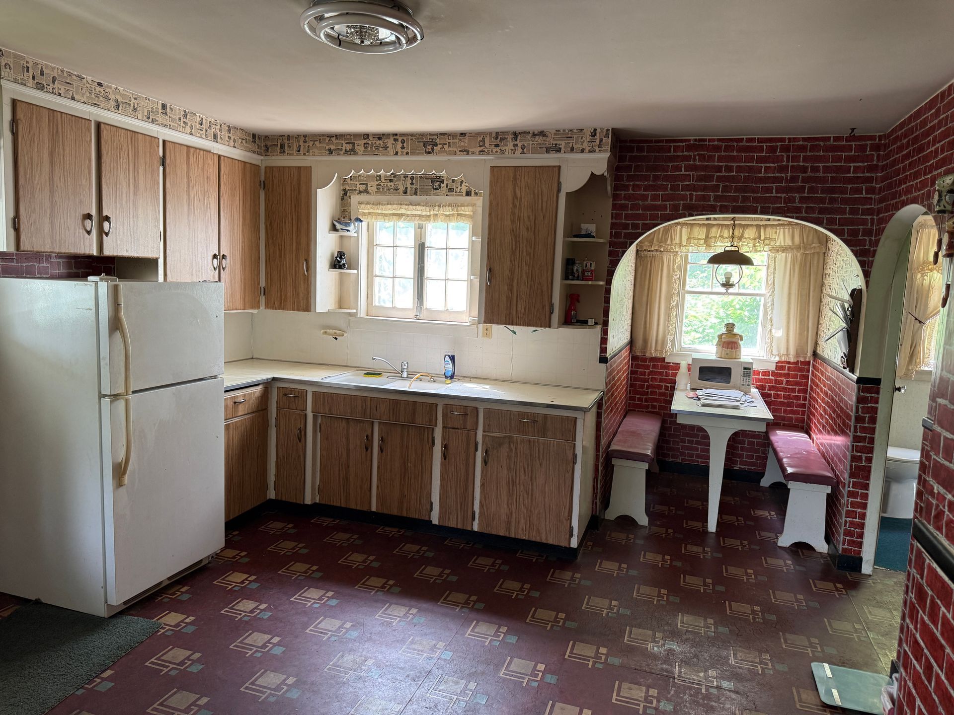 Small kitchen with wood cabinets, white fridge, sink window, and a dining nook by brick wall.