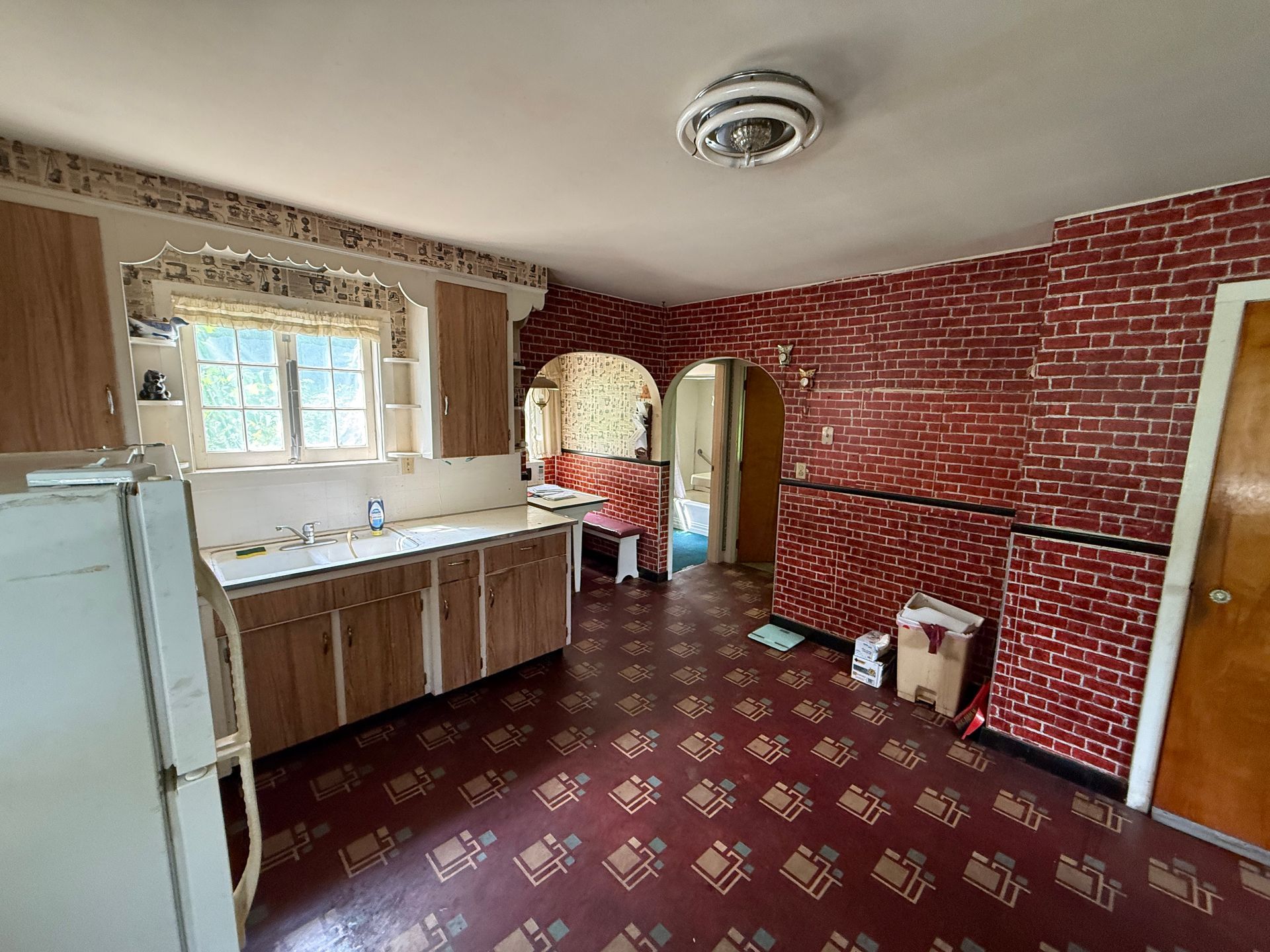 kitchen with brick walls, white fridge, wooden cabinets, patterned red floor, and archway to another room