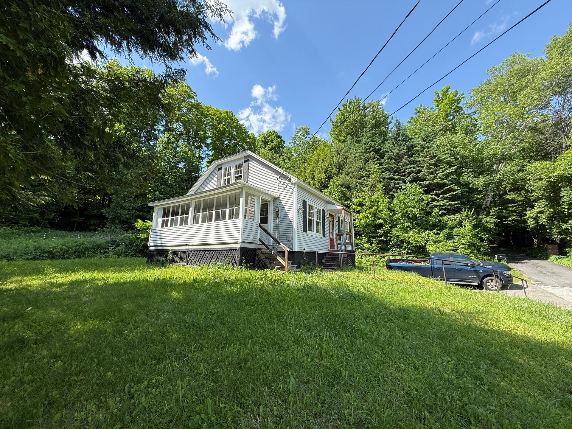 White house on a grassy hillside beside trees and power lines under a blue sky.