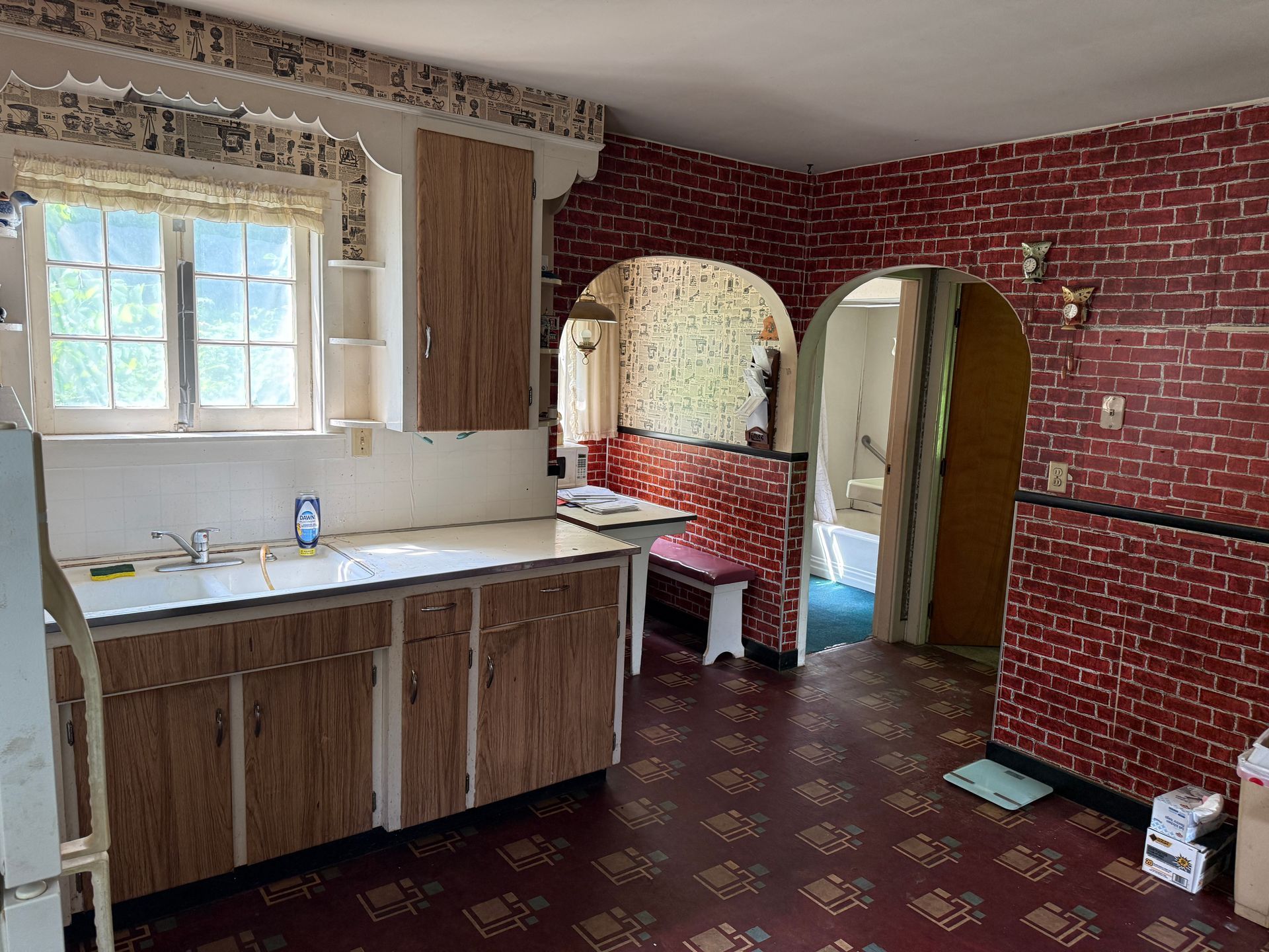 Small kitchen with brick-pattern wallpaper, wooden cabinets, window, and arched doorway to another room
