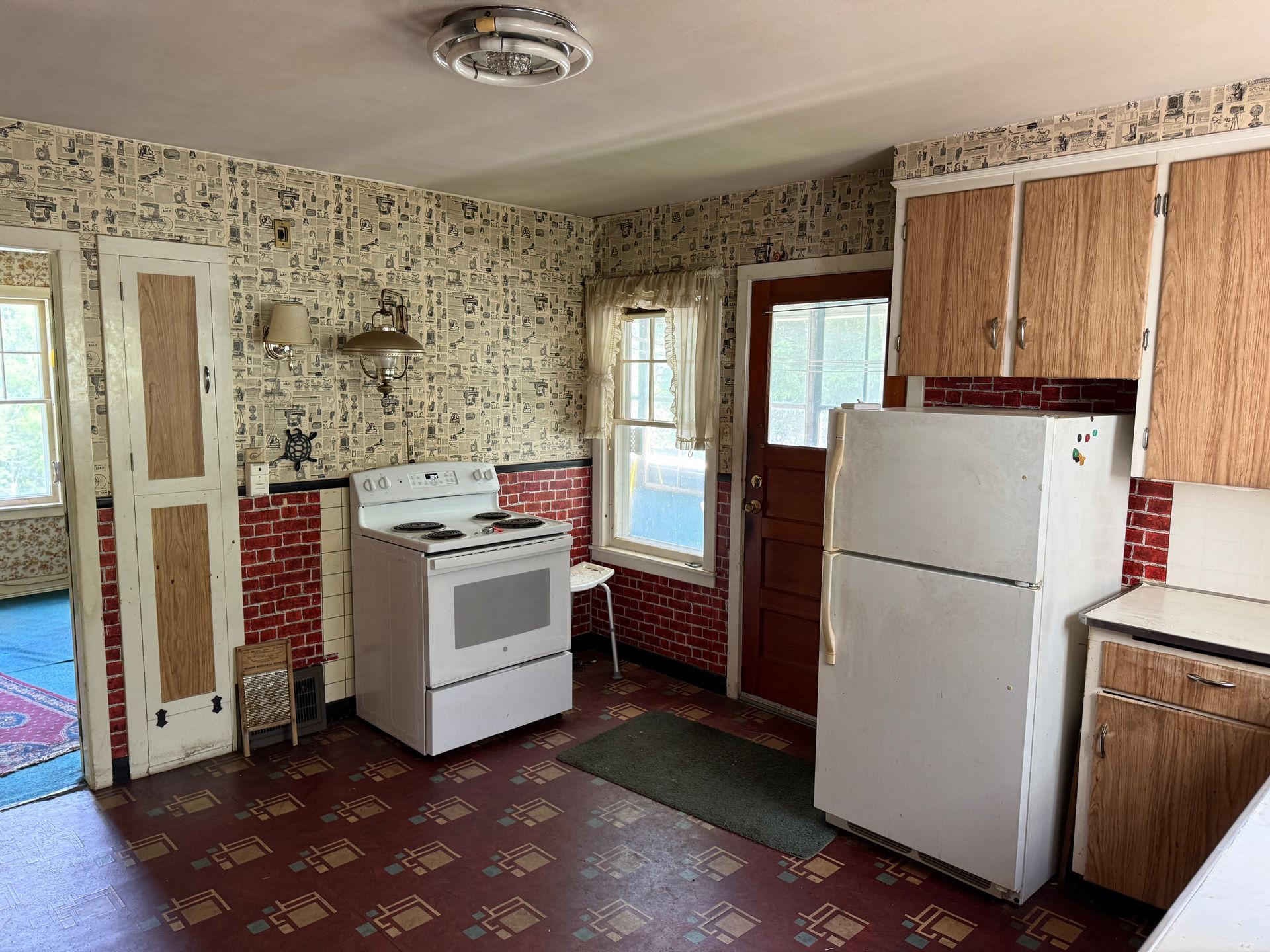 Small vintage kitchen with white stove and refrigerator, wood cabinets, patterned wallpaper, and red tile flooring.