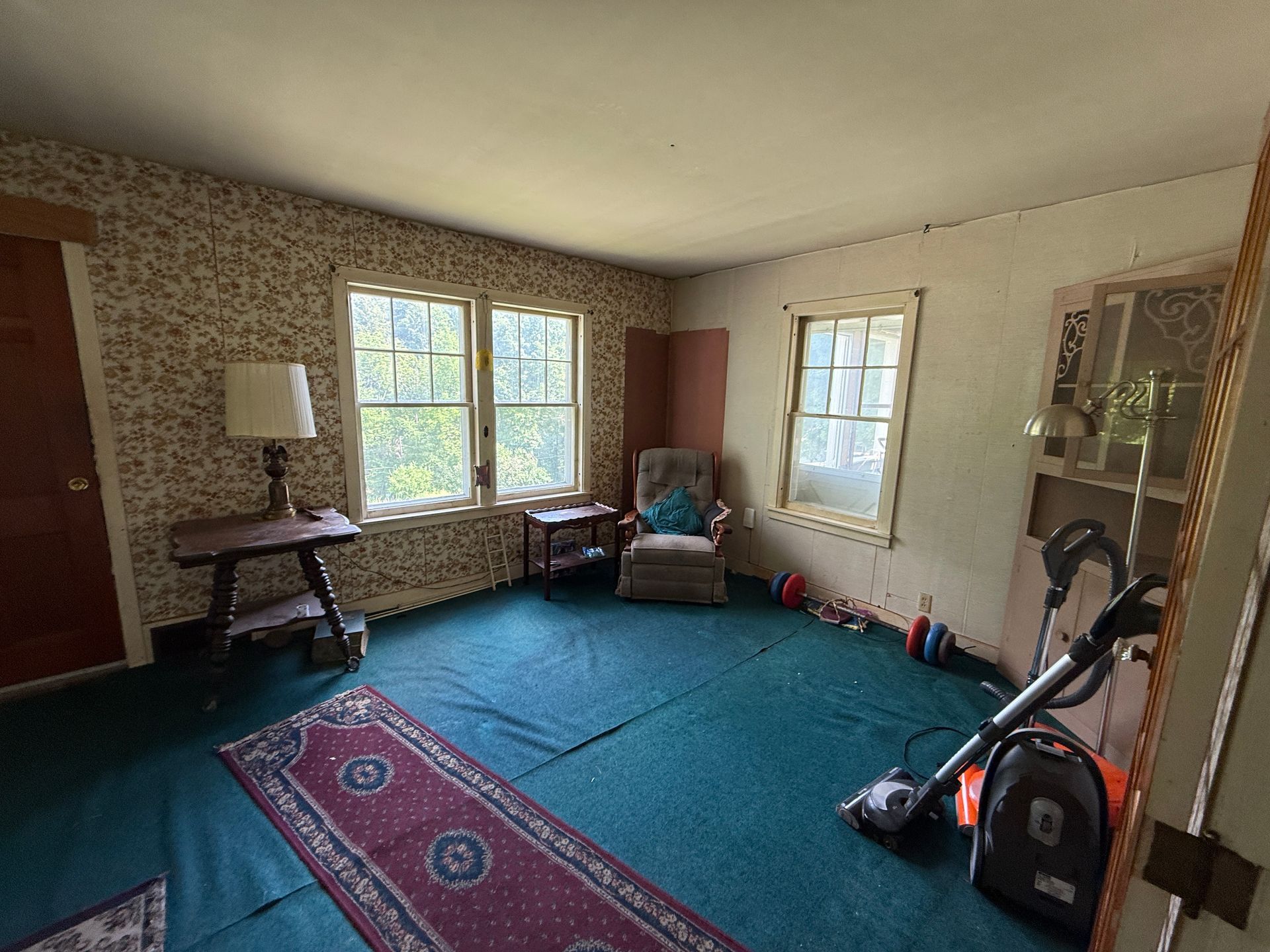 Cozy living room with teal carpet, floral rug, windows, armchair, lamp, and a vacuum cleaner.