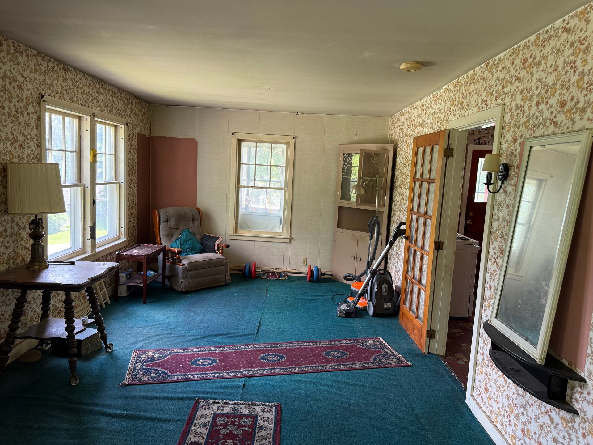 Empty living room with floral wallpaper, blue carpet, large windows, and a rug in the center.