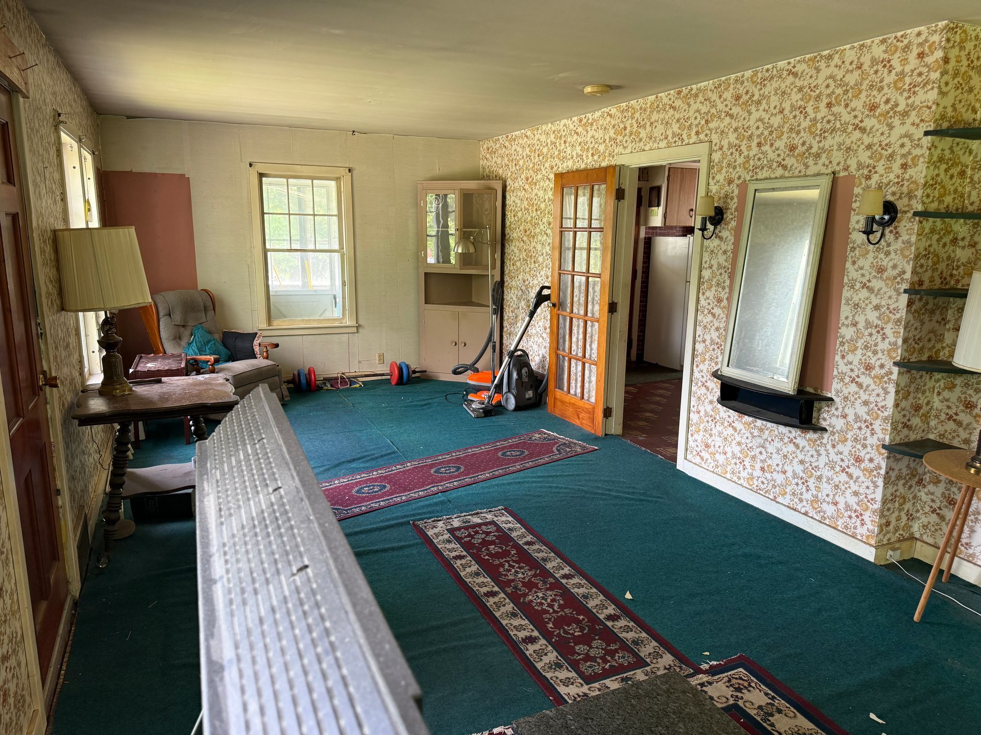 Living room with teal carpet, patterned wallpaper, windows, a lamp, and a fireplace opening
