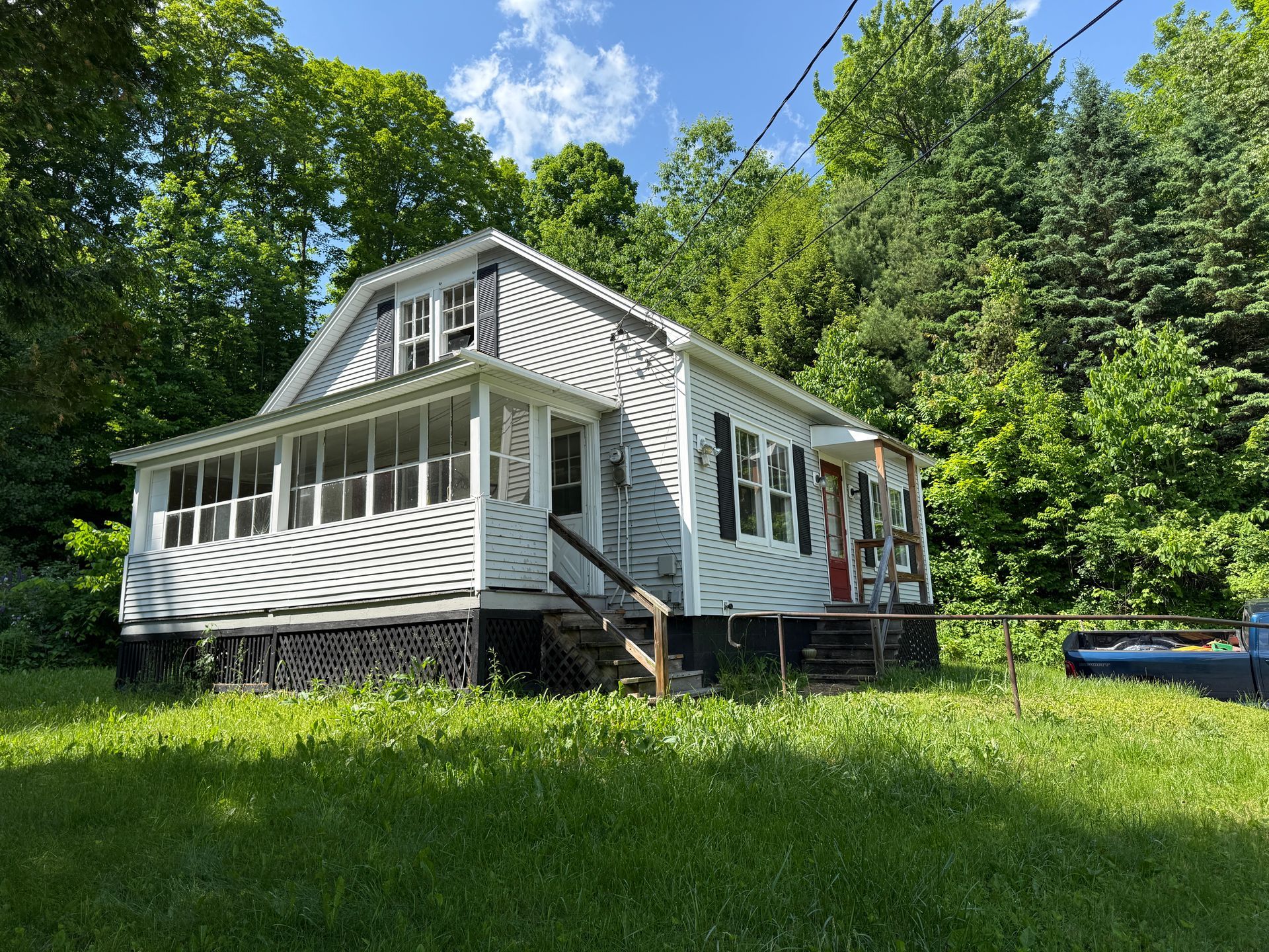 Small white house with porch in a grassy clearing surrounded by trees under a blue sky