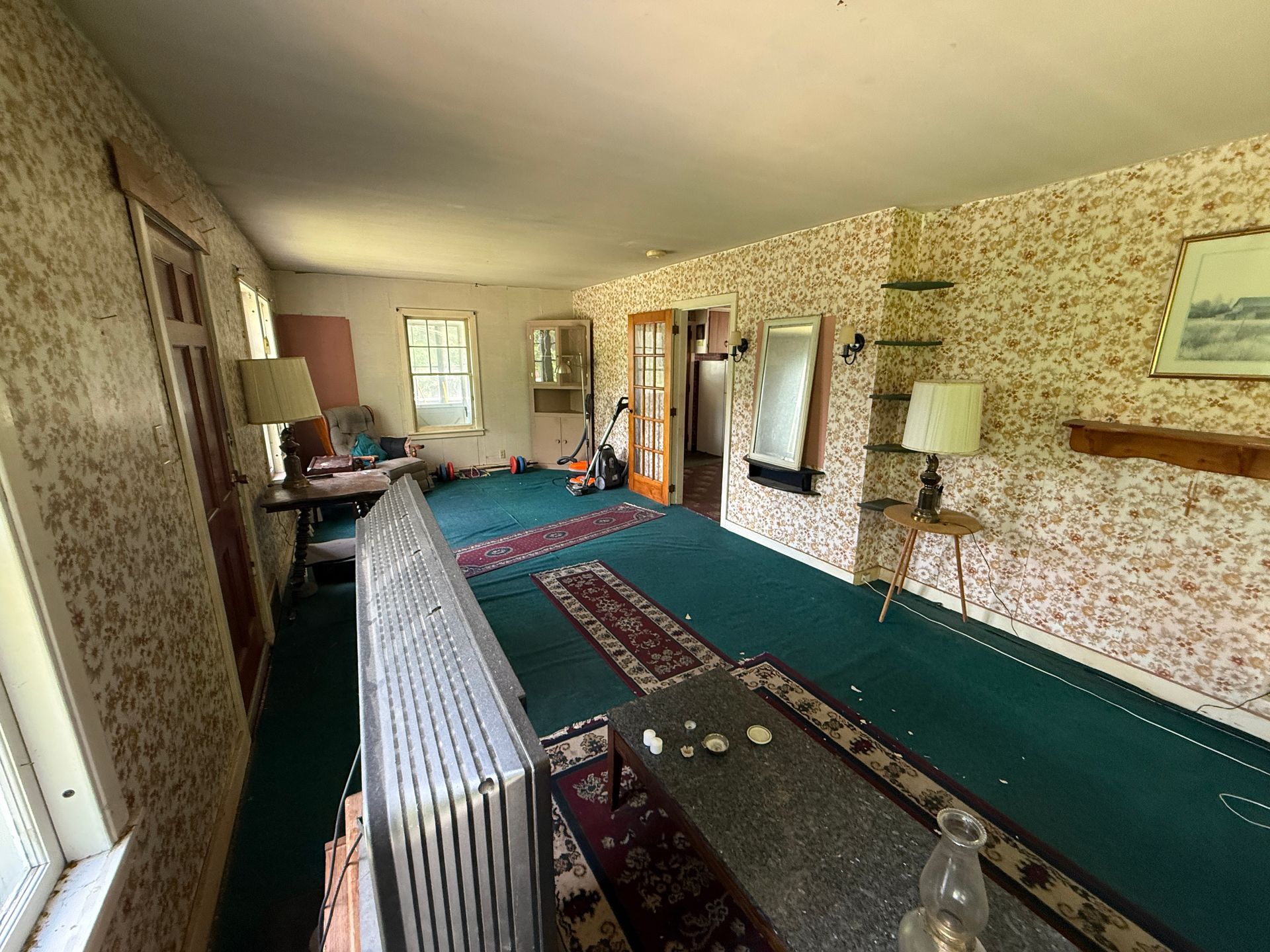 Long living room with patterned wallpaper, green carpet, radiator, and a window at the far end