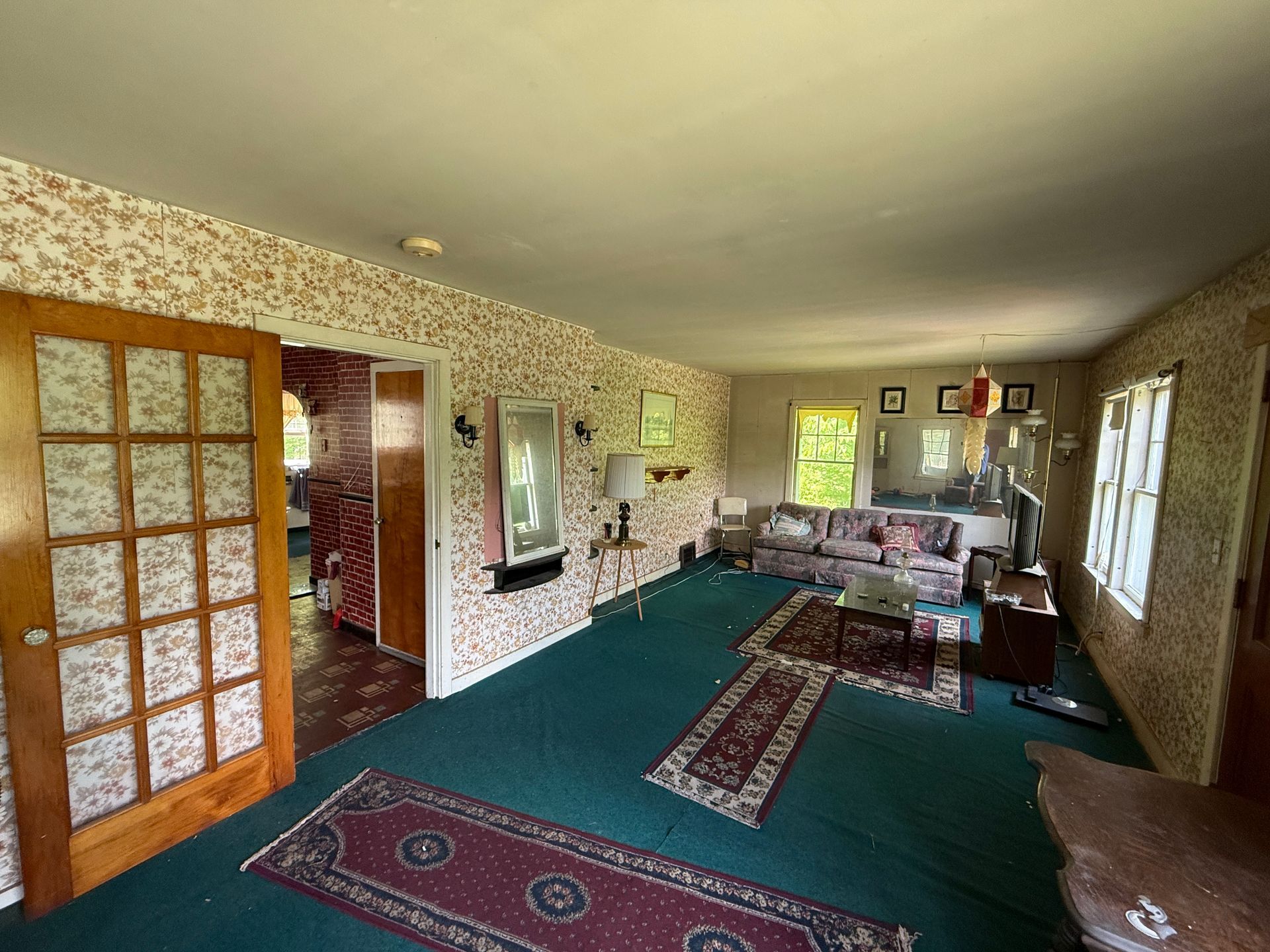 Spacious living room with green carpet, patterned wallpaper, wooden French doors, and a sofa by large windows.