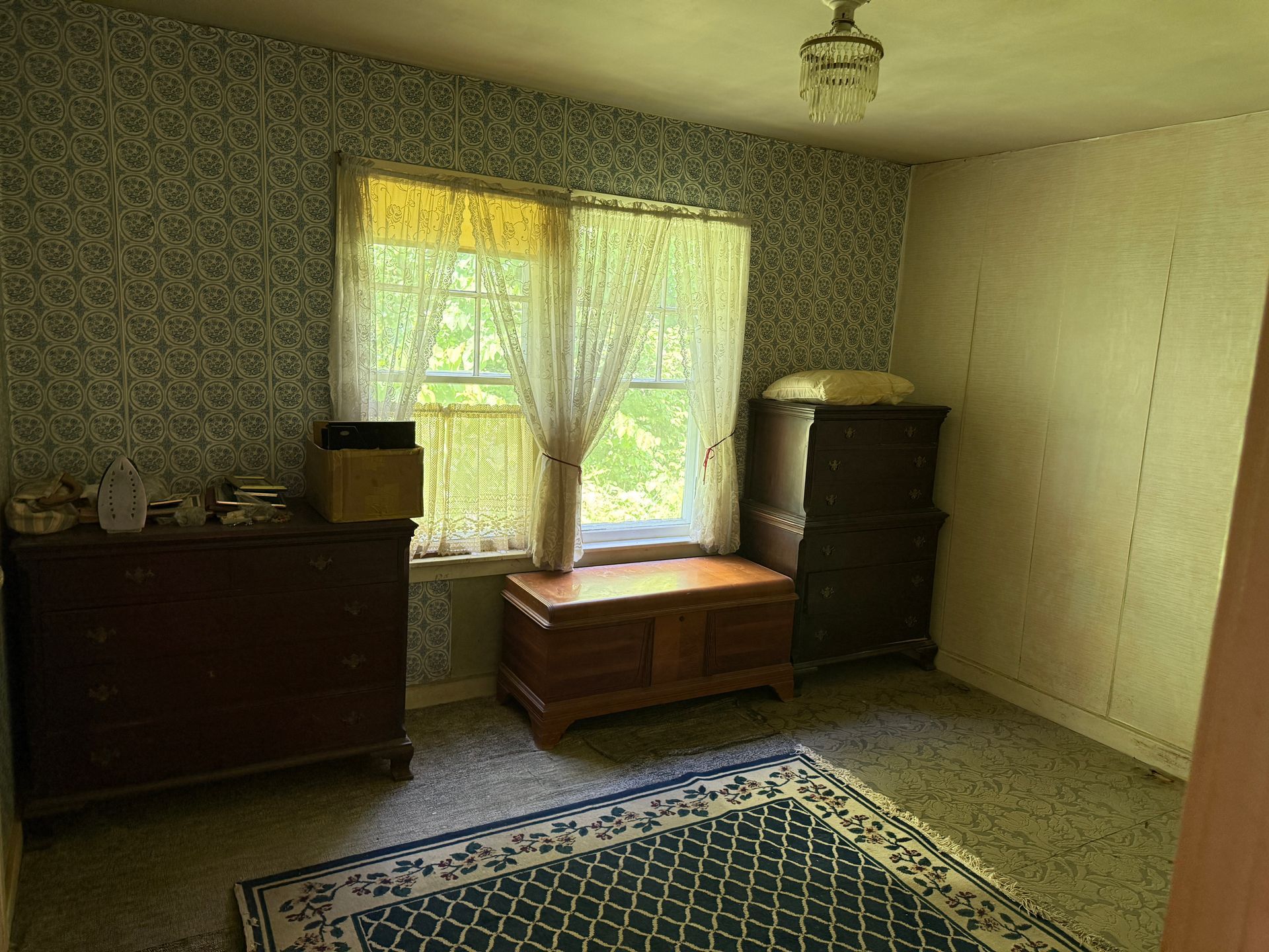 Small furnished room with window, dresser, wooden chest, and blue patterned rug.