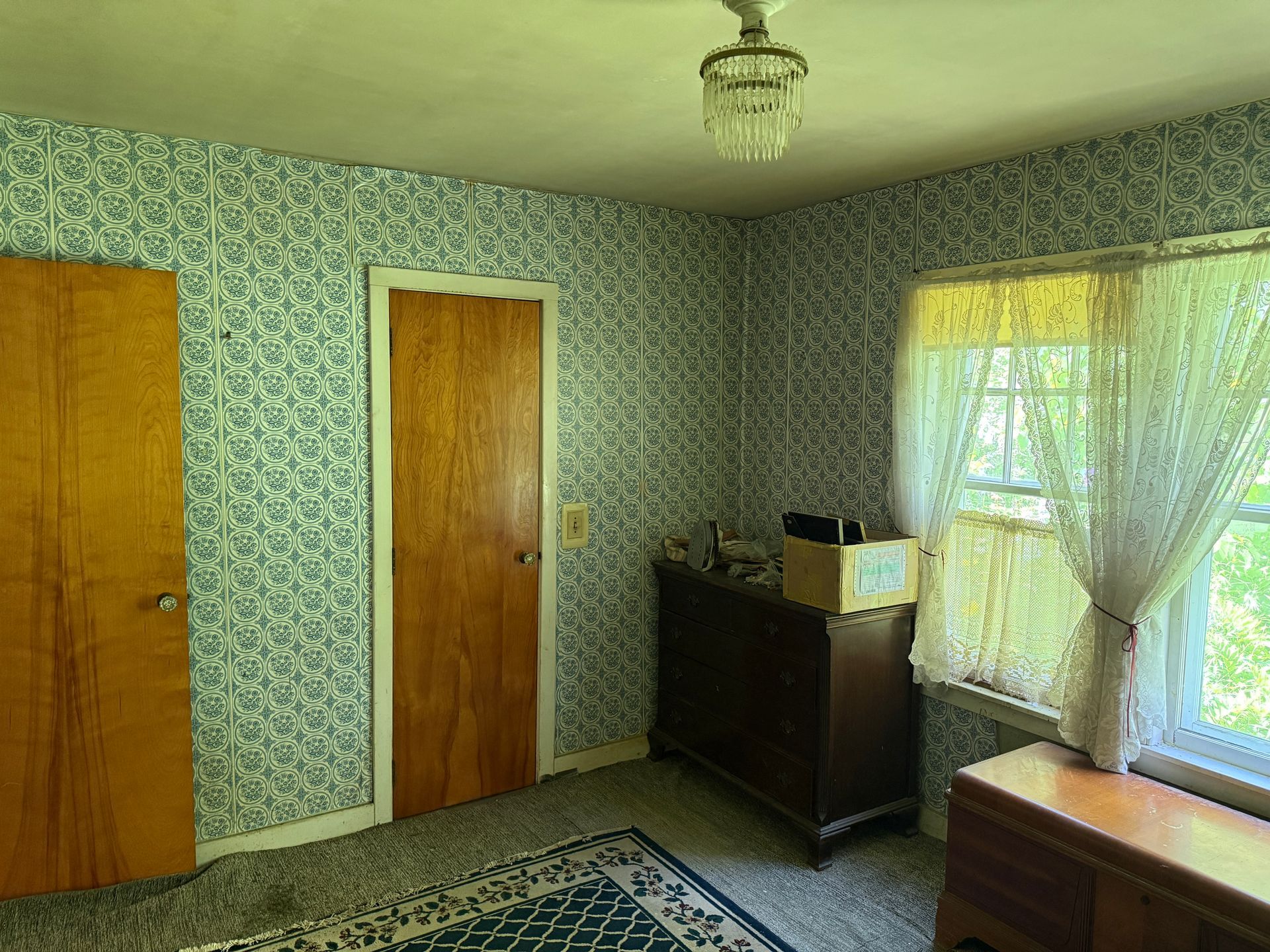 Empty bedroom with patterned wallpaper, two wooden doors, dresser, and sunlit window with sheer curtains.