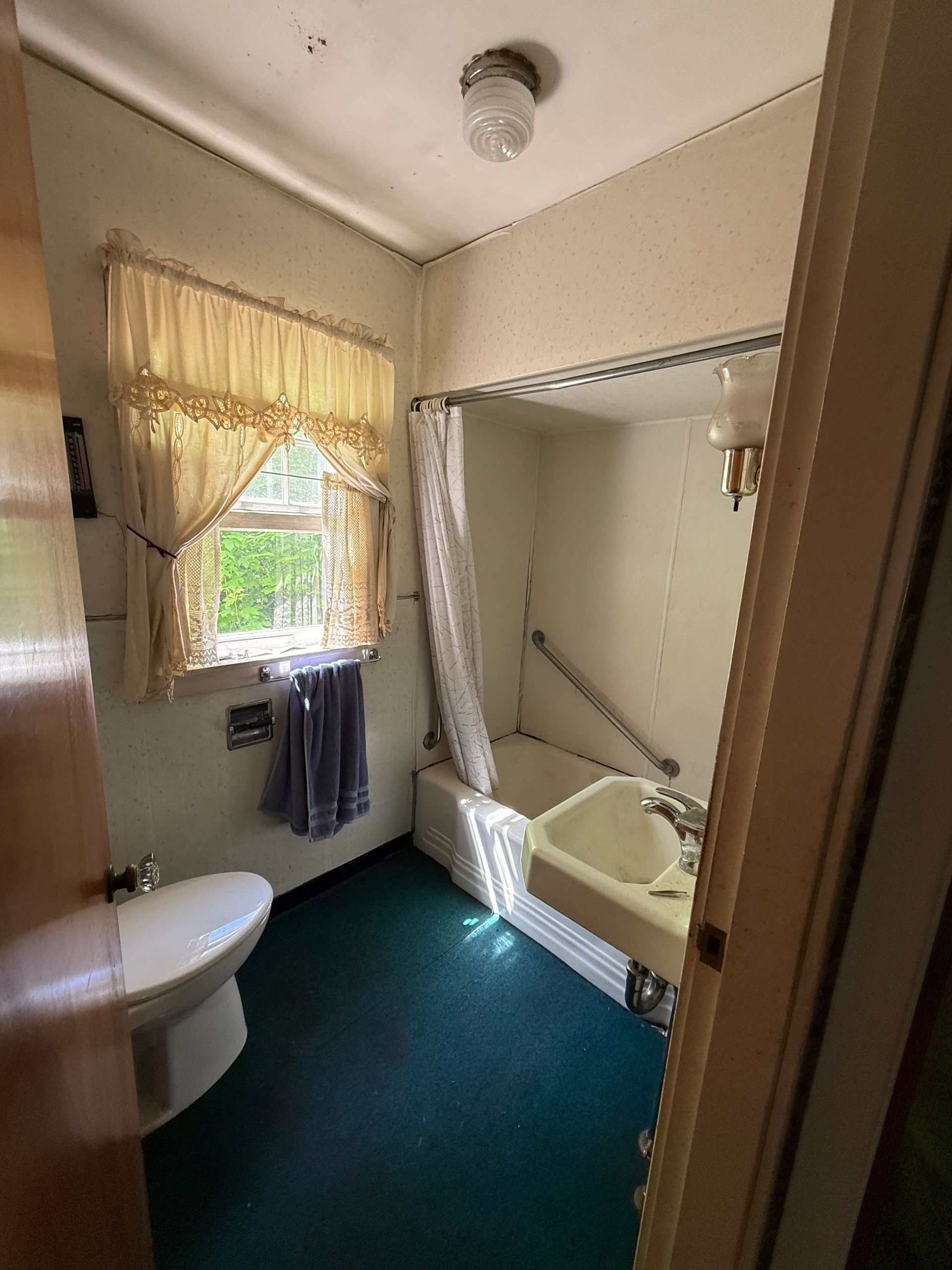 Small bathroom with shower-tub, toilet, window with curtains, and blue carpeted floor.