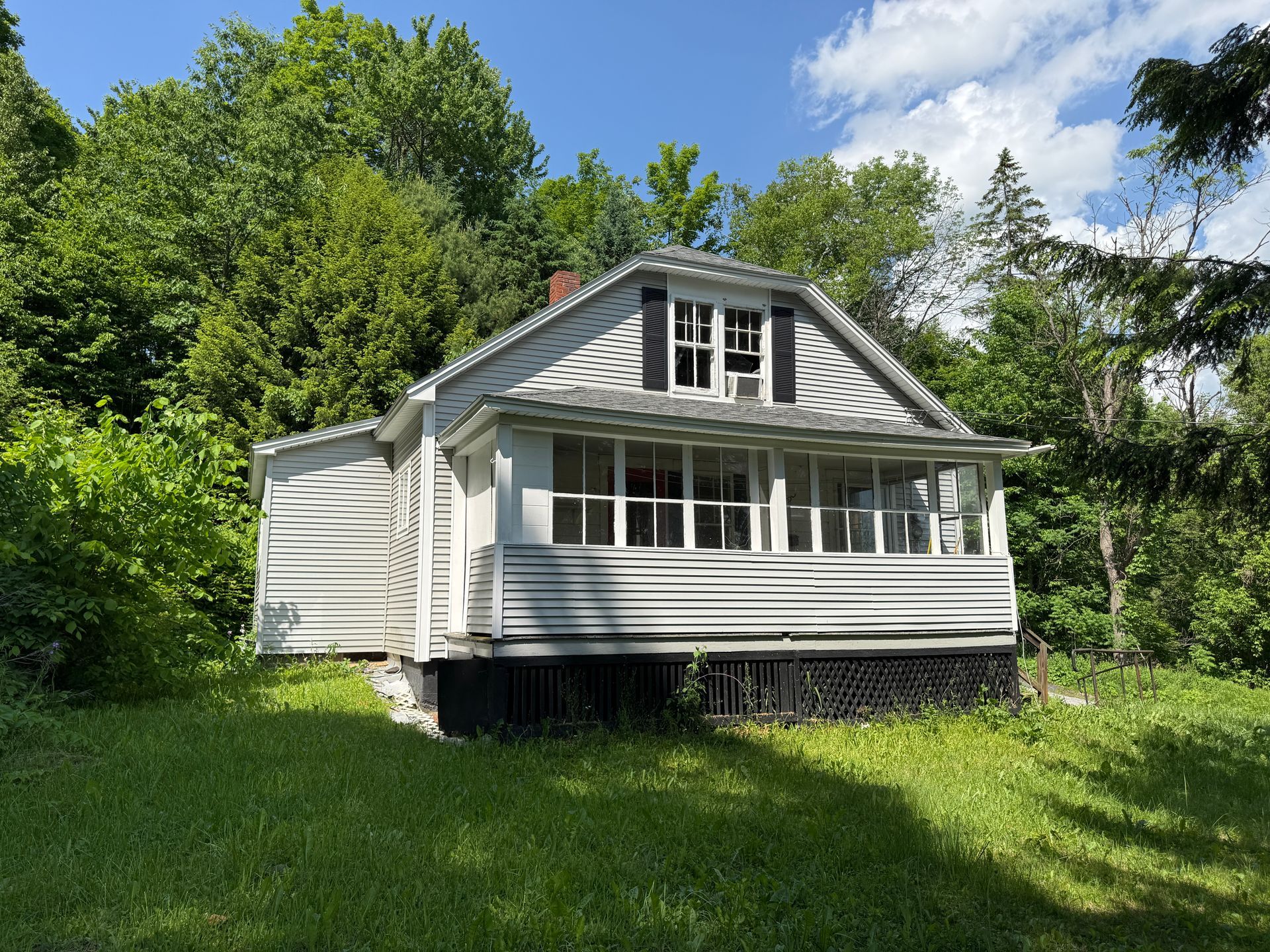 Small white cottage with a porch and dormer, surrounded by green grass and trees under a blue sky.