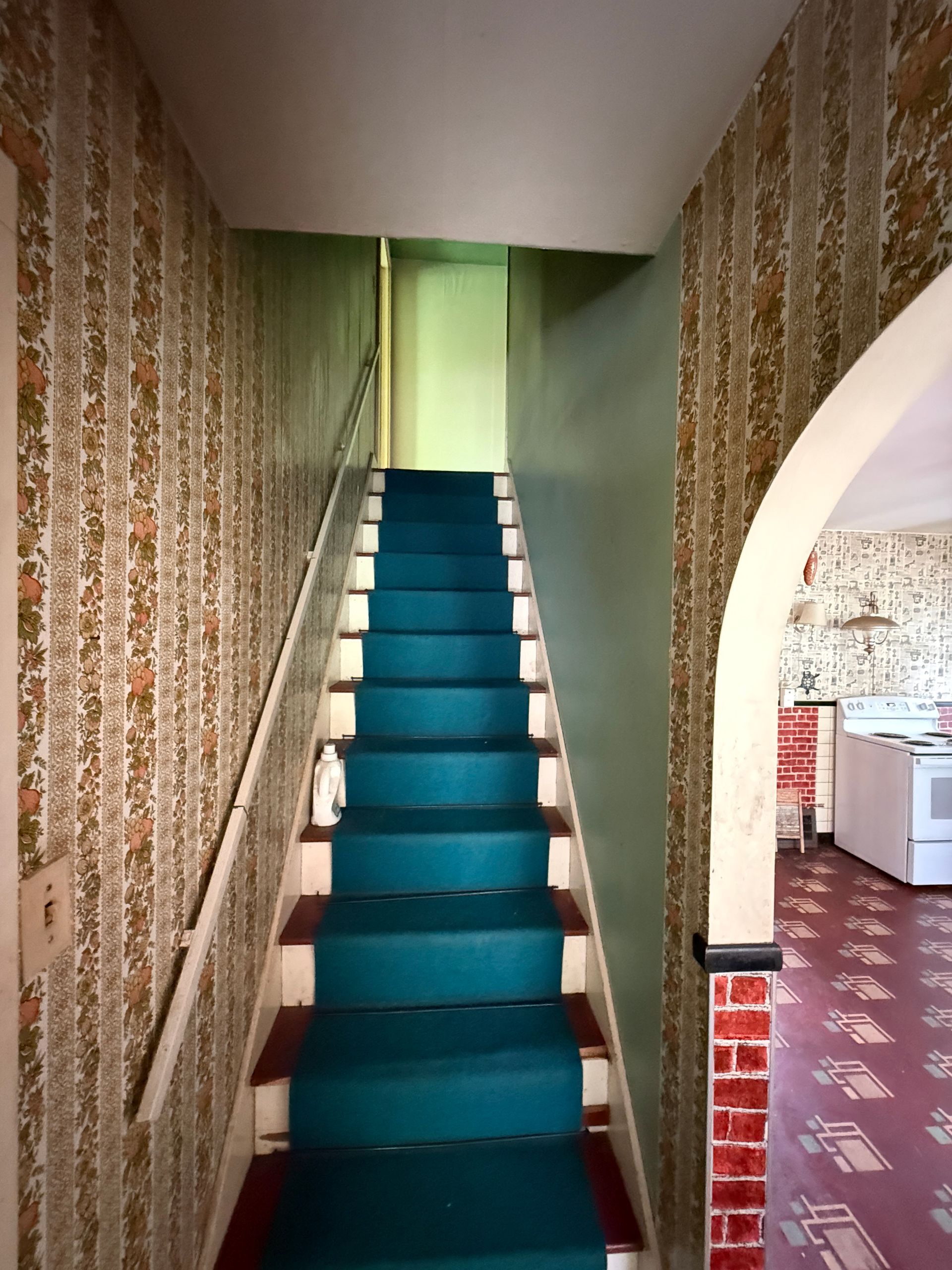 Narrow staircase with blue carpet in a patterned hallway, leading to a bright doorway.