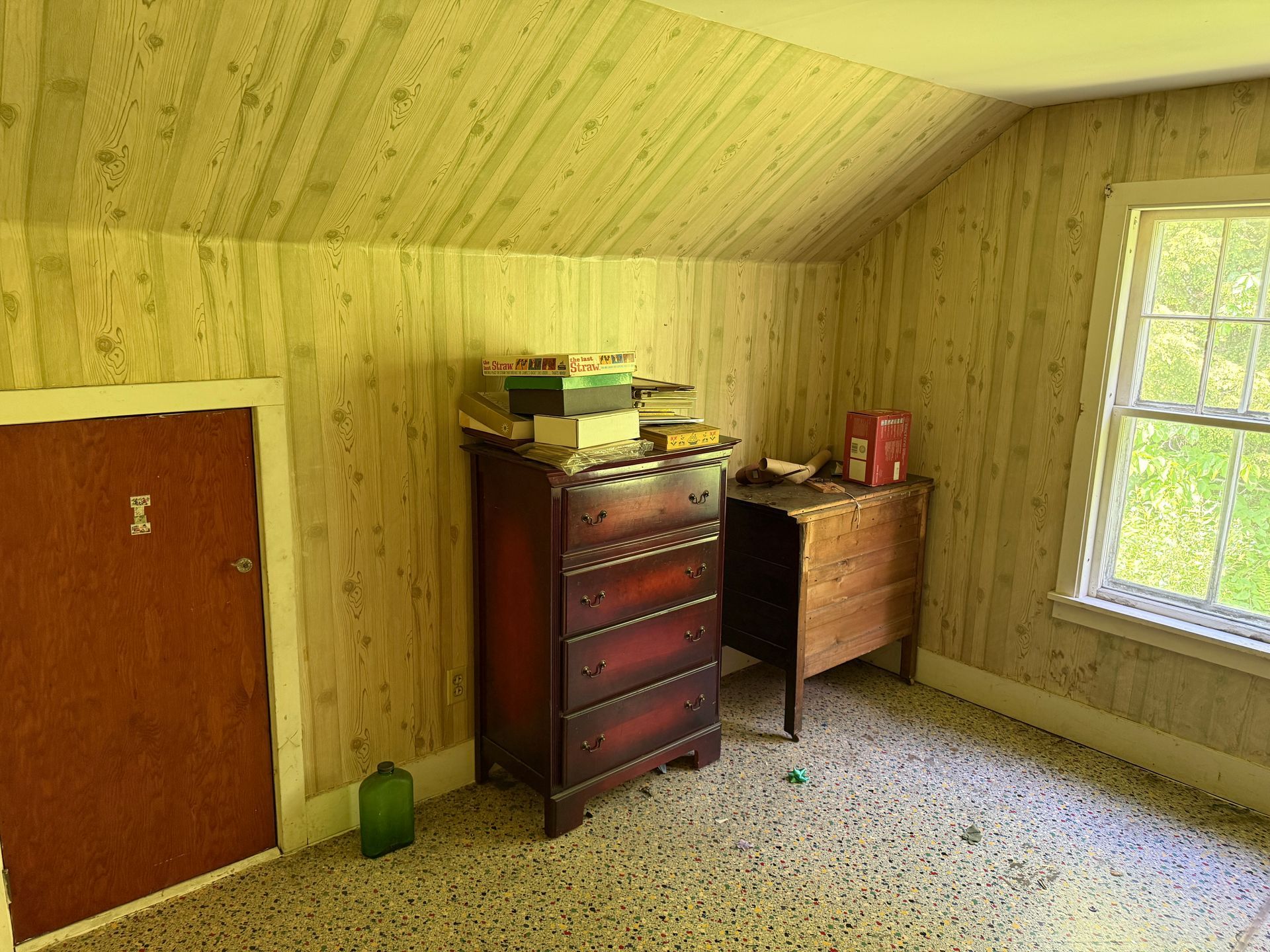 Small attic bedroom with wood-paneled walls, two dressers, a door, and a window with green view.