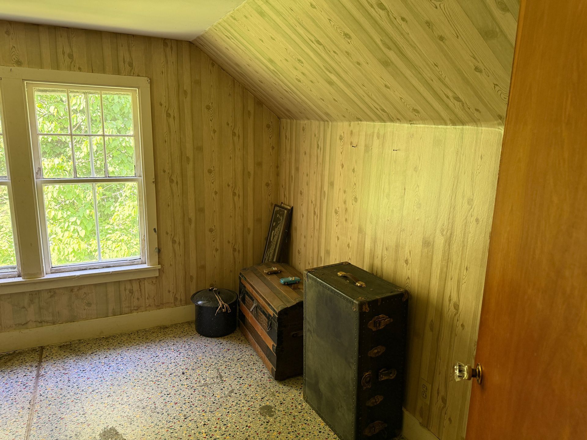 Small wood-paneled attic room with a window, carpet, and a few boxes and furniture pieces.