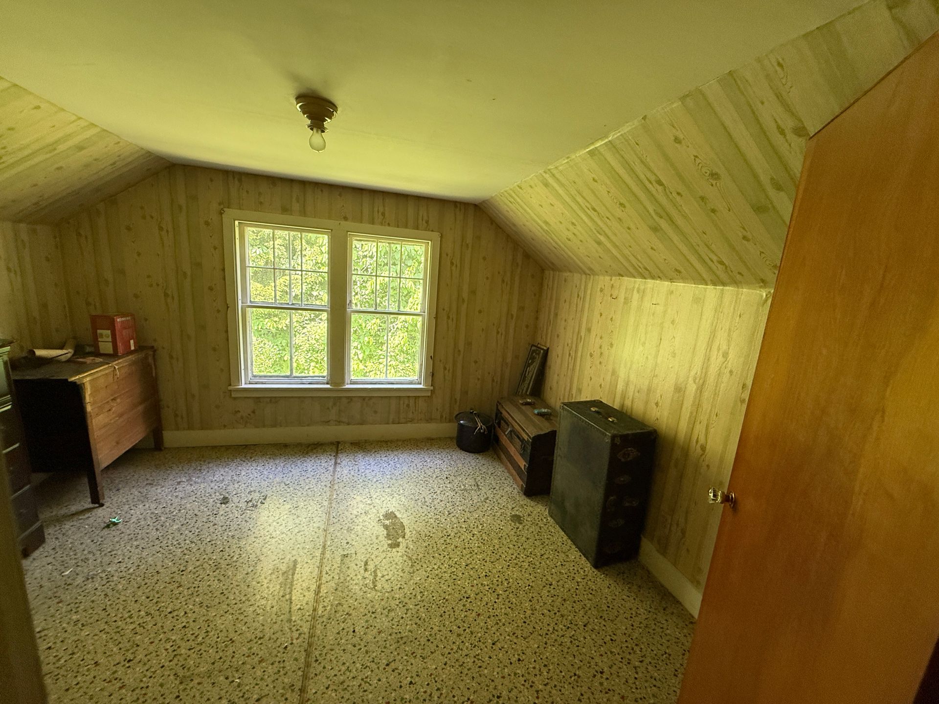 Empty attic room with wood-paneled walls, slanted ceiling, and a window at the far end.