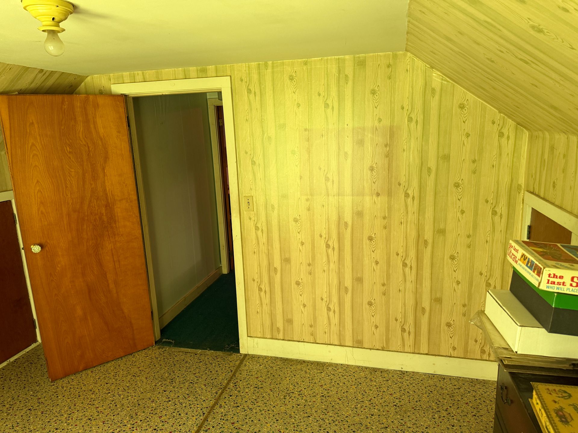 Small attic room with slanted ceiling, open wooden door, patterned wallpaper, and a desk with boxes.