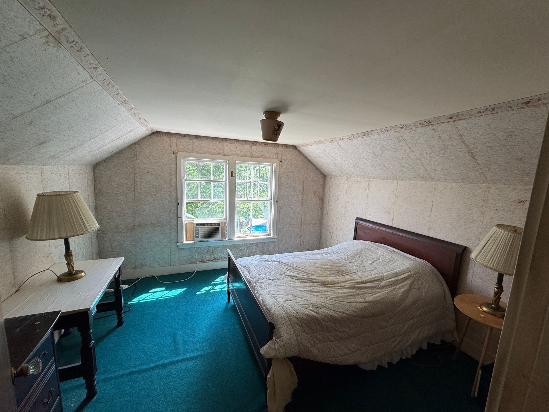 Cozy attic bedroom with bed, two lamps, a desk, and a window with daylight and green rug.