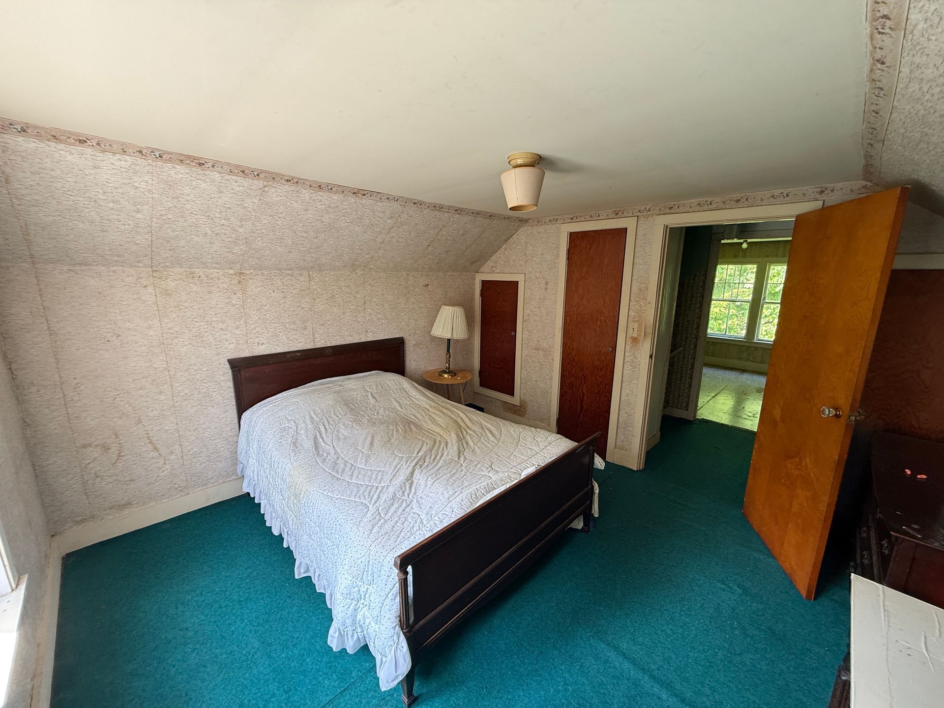 Bedroom with double bed, teal carpet, wooden doors, and a window letting in daylight.