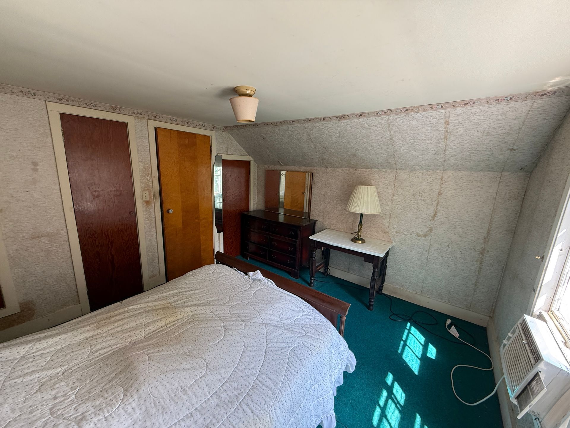 Small bedroom with a bed, dresser, desk, and two doors, lit by sunlight from a window.