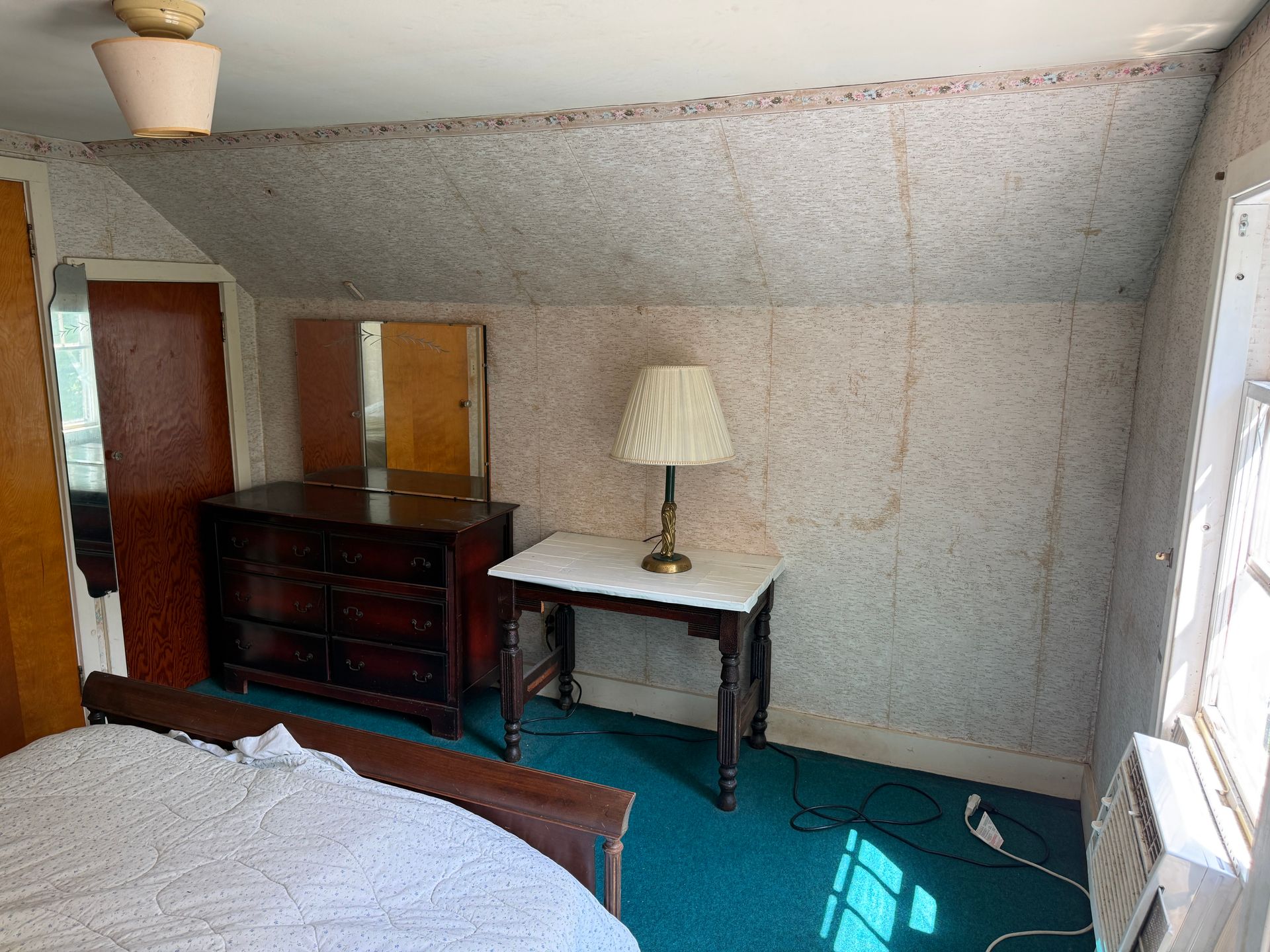 Bedroom with slanted ceiling, bed, dresser, table lamp, teal rug, and a window air conditioner.