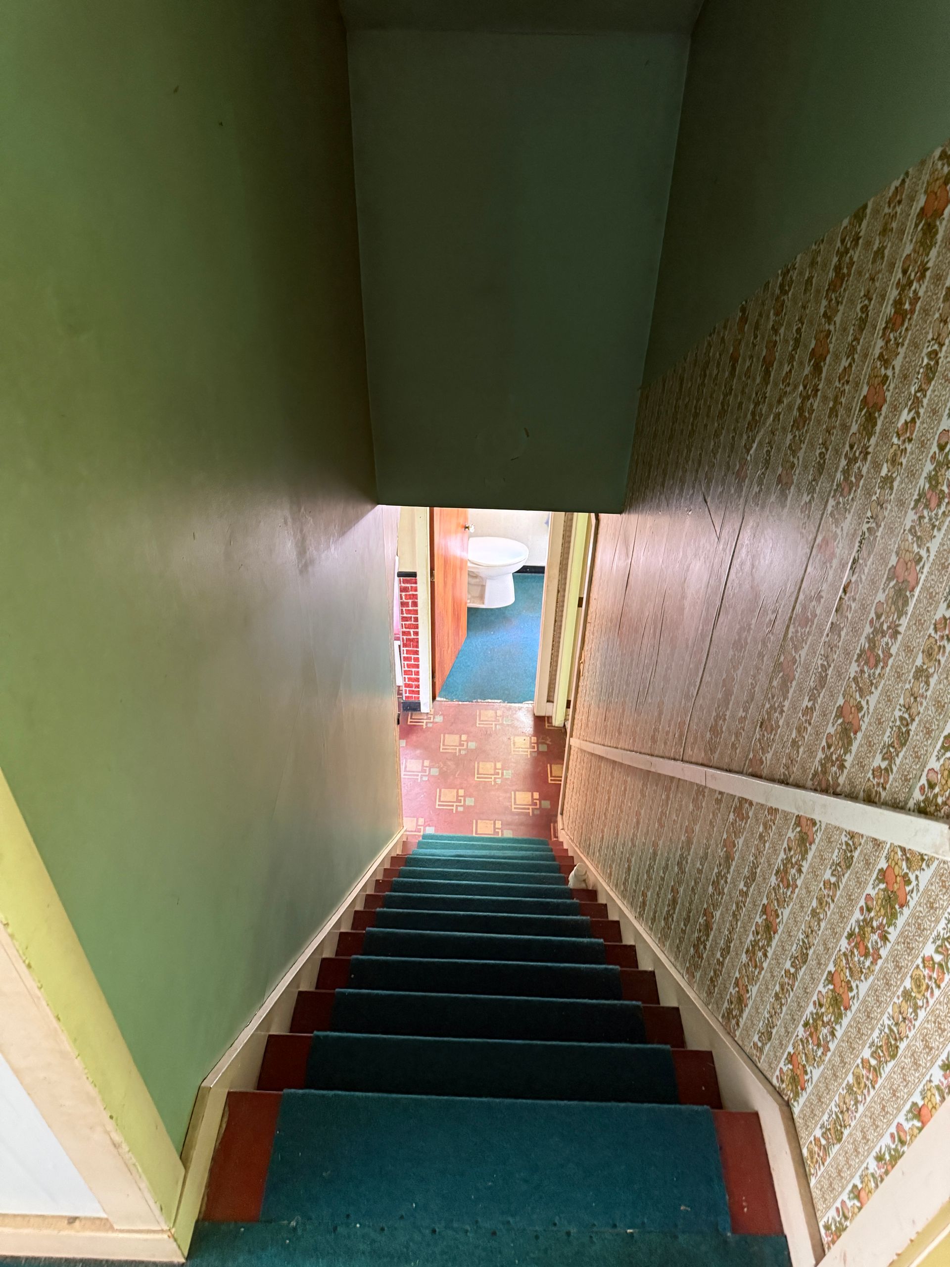 Narrow stairwell with green walls, red steps, and patterned wallpaper leading to a doorway.