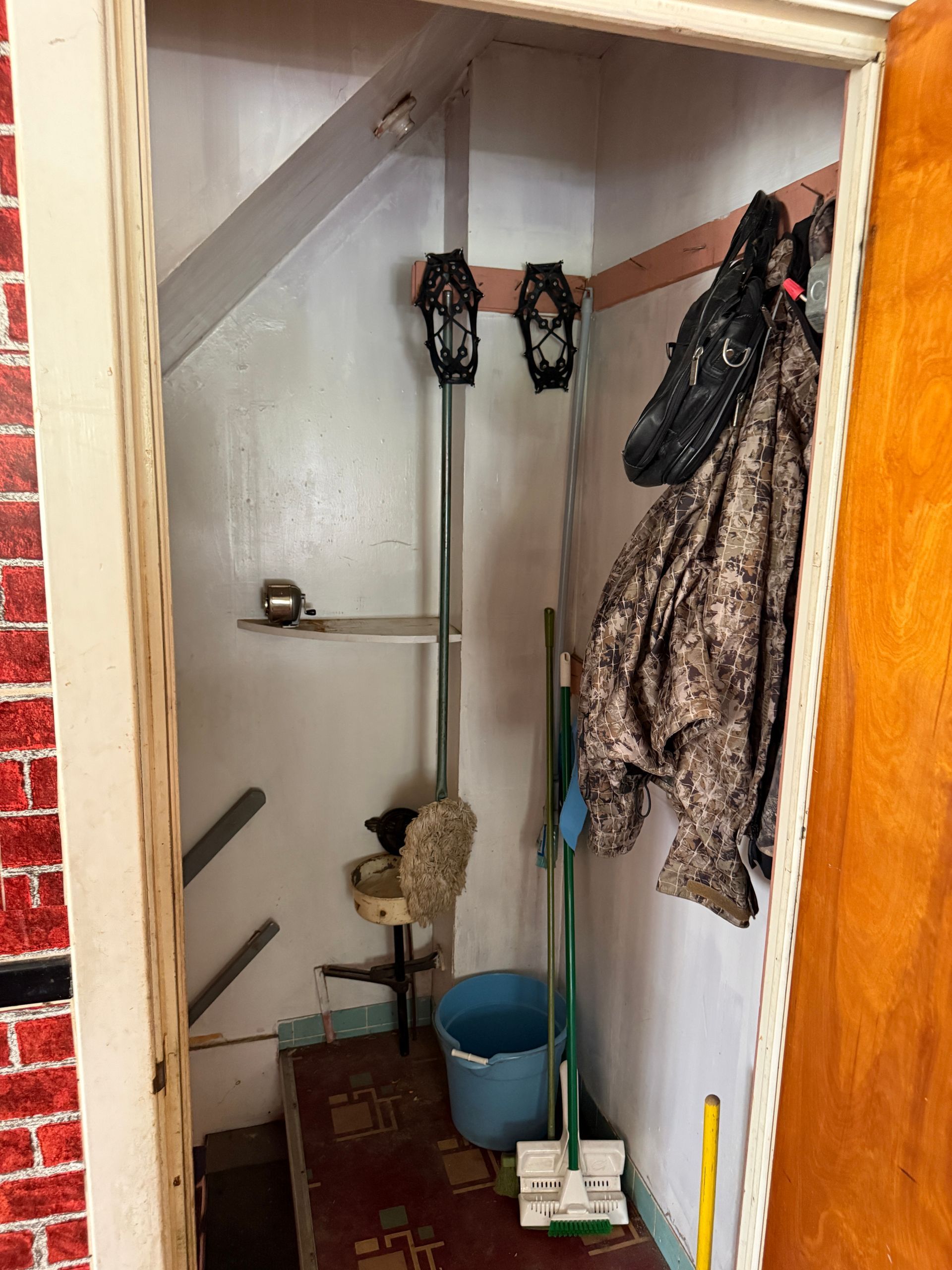 Small utility closet with hanging clothes, cleaning supplies, and a blue bucket on the floor.