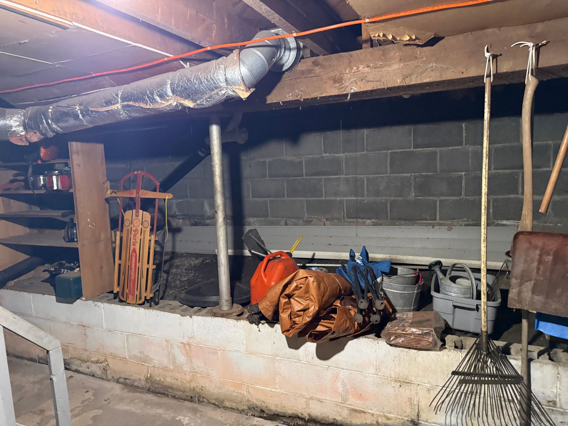 Basement crawlspace with cinderblock wall, pipes, tools, and stored materials under a low ceiling