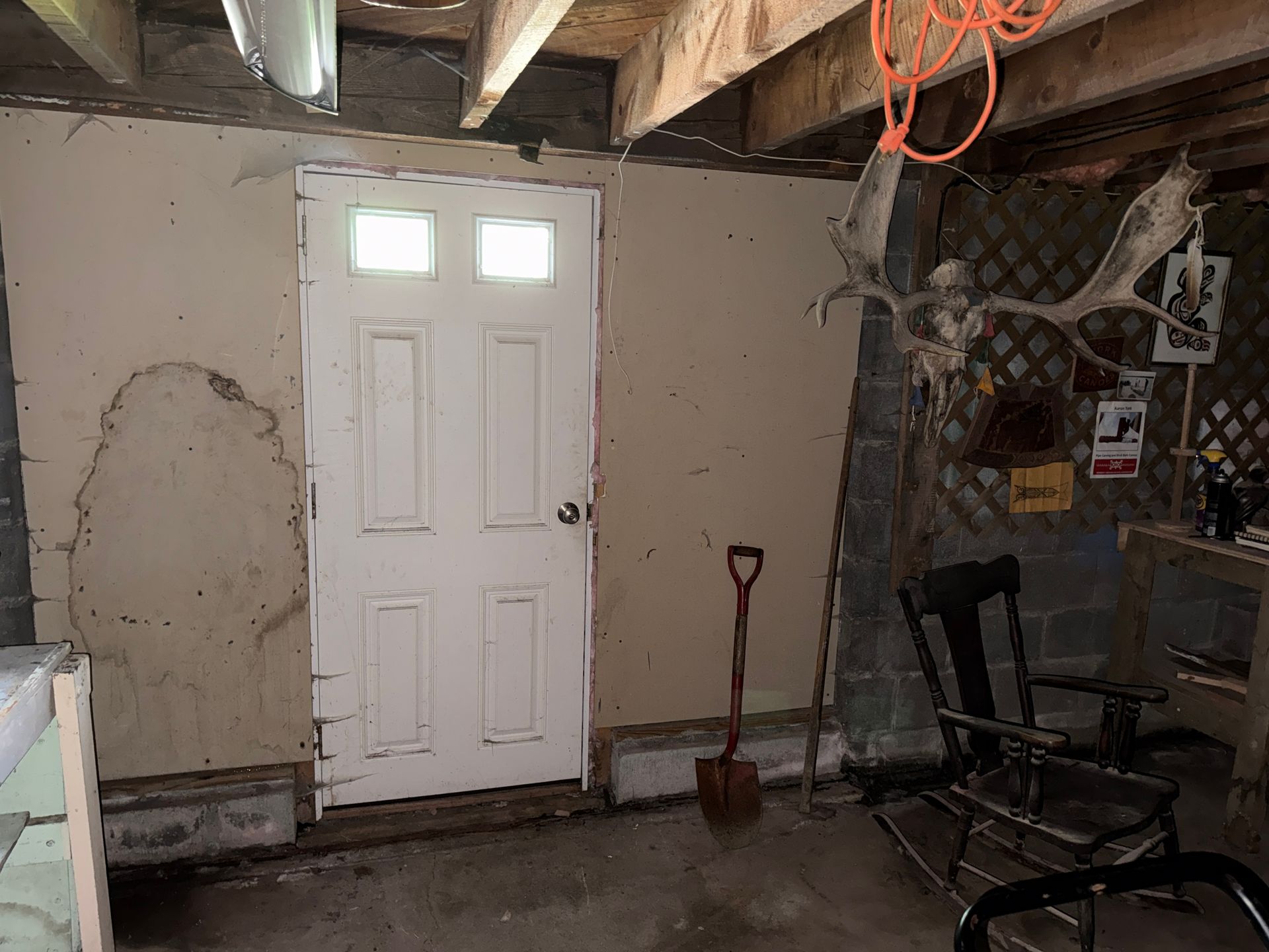 Basement/garage interior with white door, exposed ceiling beams, shovel, and stacked lawn chairs.