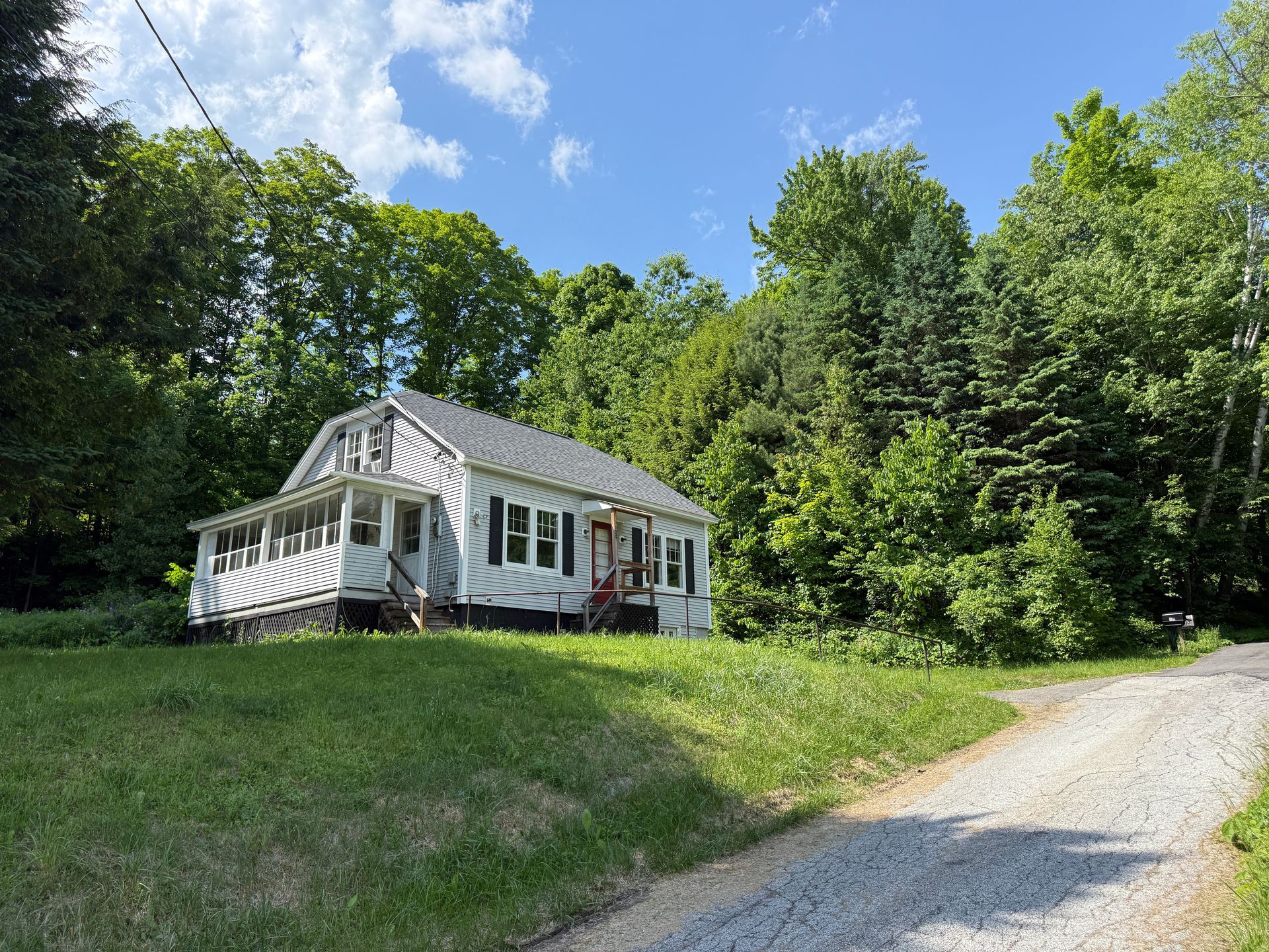 Small white house on a grassy hill beside a gravel road, surrounded by trees under a blue sky.