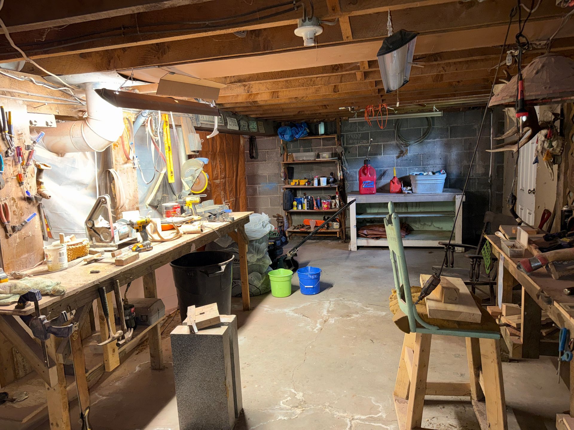 basement workshop with workbenches, tools, shelves, buckets, and a wooden chair under exposed beams.