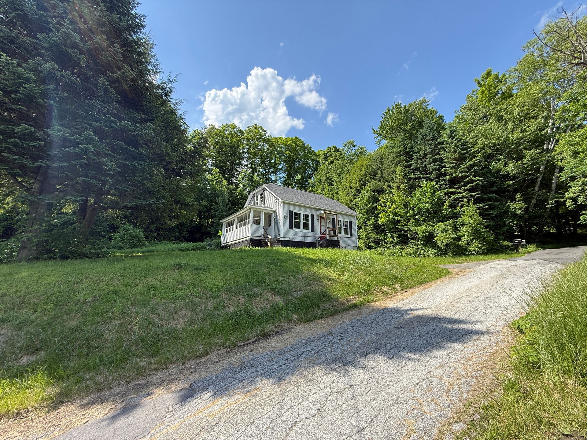 Small white house with a gravel driveway, surrounded by green trees under a blue sky.