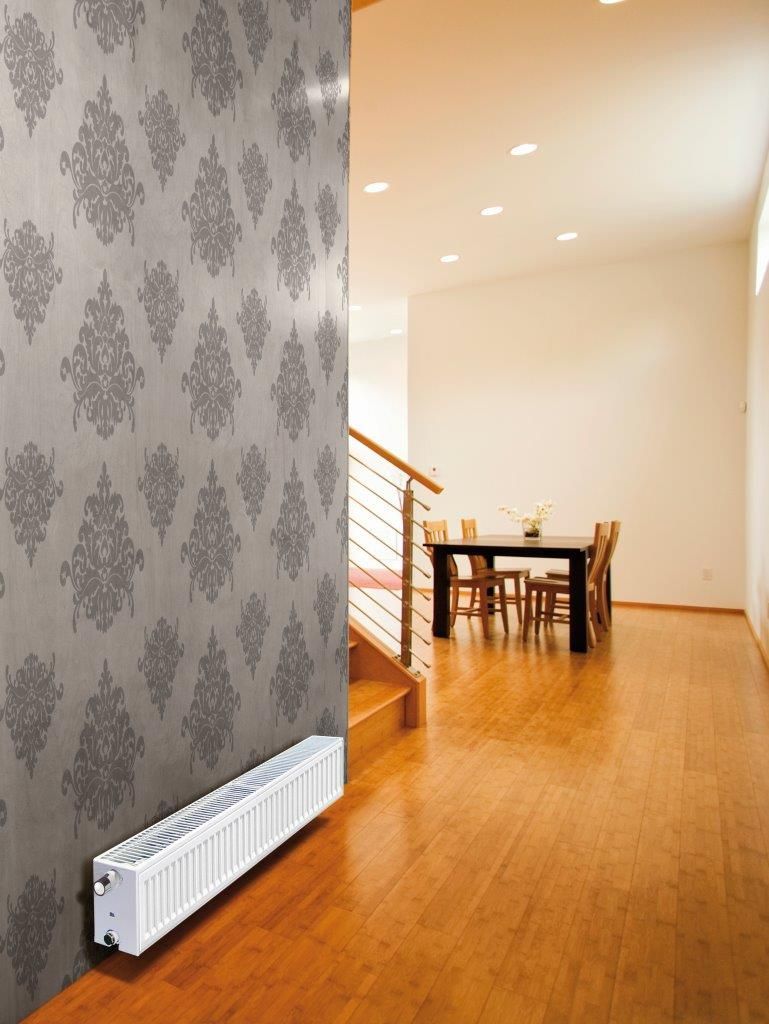 A white radiator mounted on a wall with gray patterned wallpaper in a hallway.