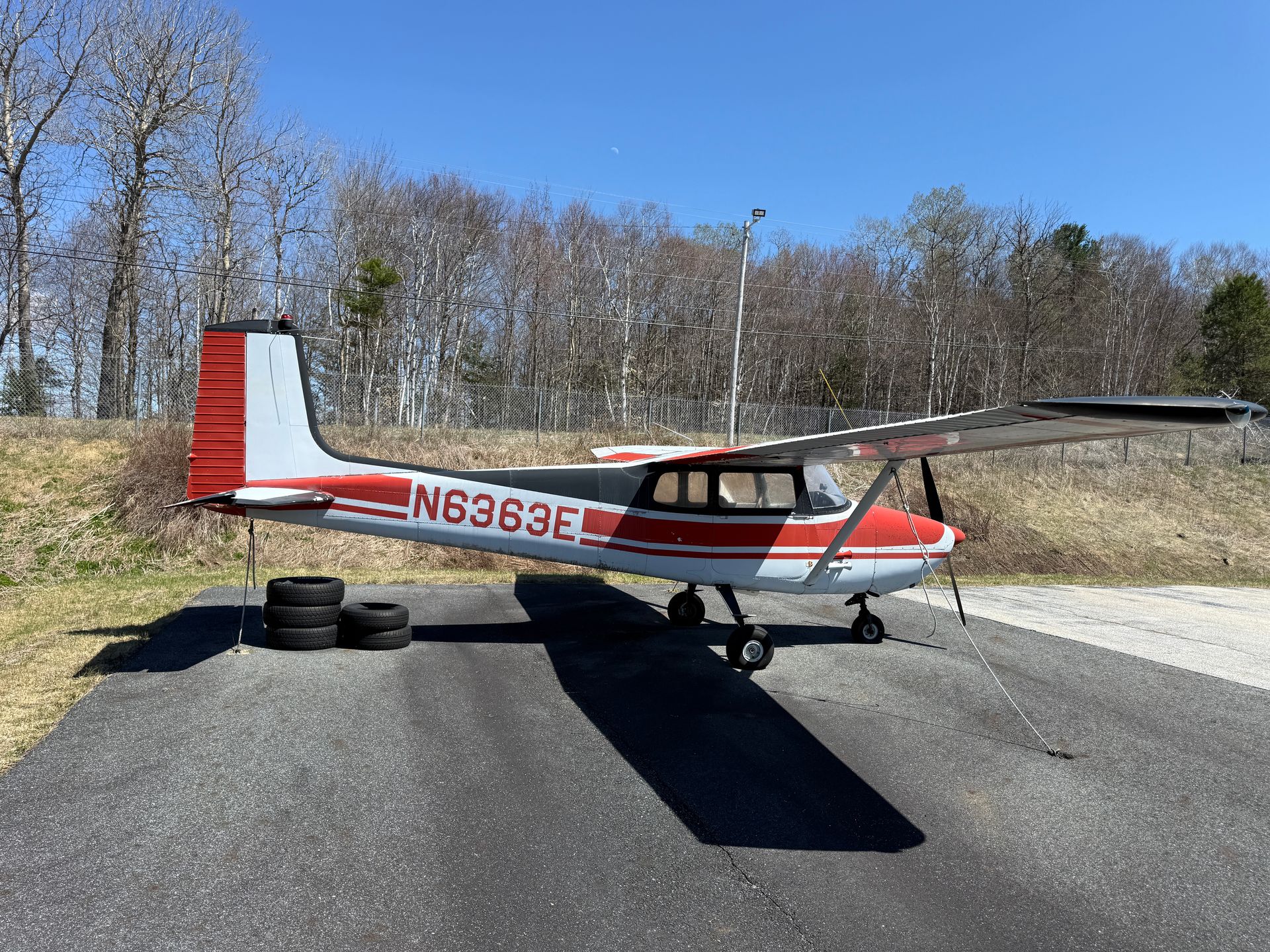 Small red-and-white airplane parked on a runway beside tires and trees under a clear blue sky