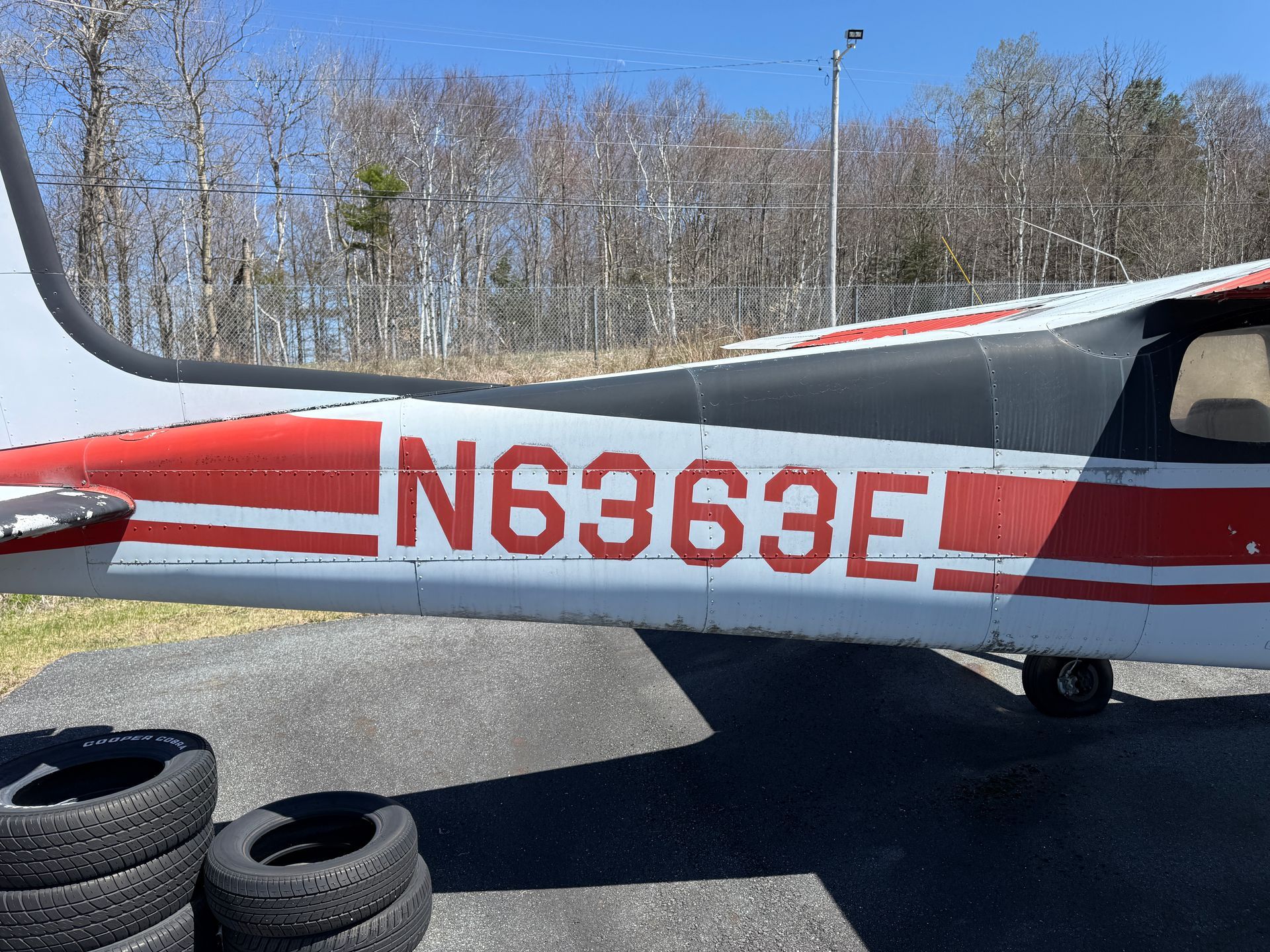 Side view of a small white airplane with red stripes and tail number N6363E on a tarmac, with trees in the background