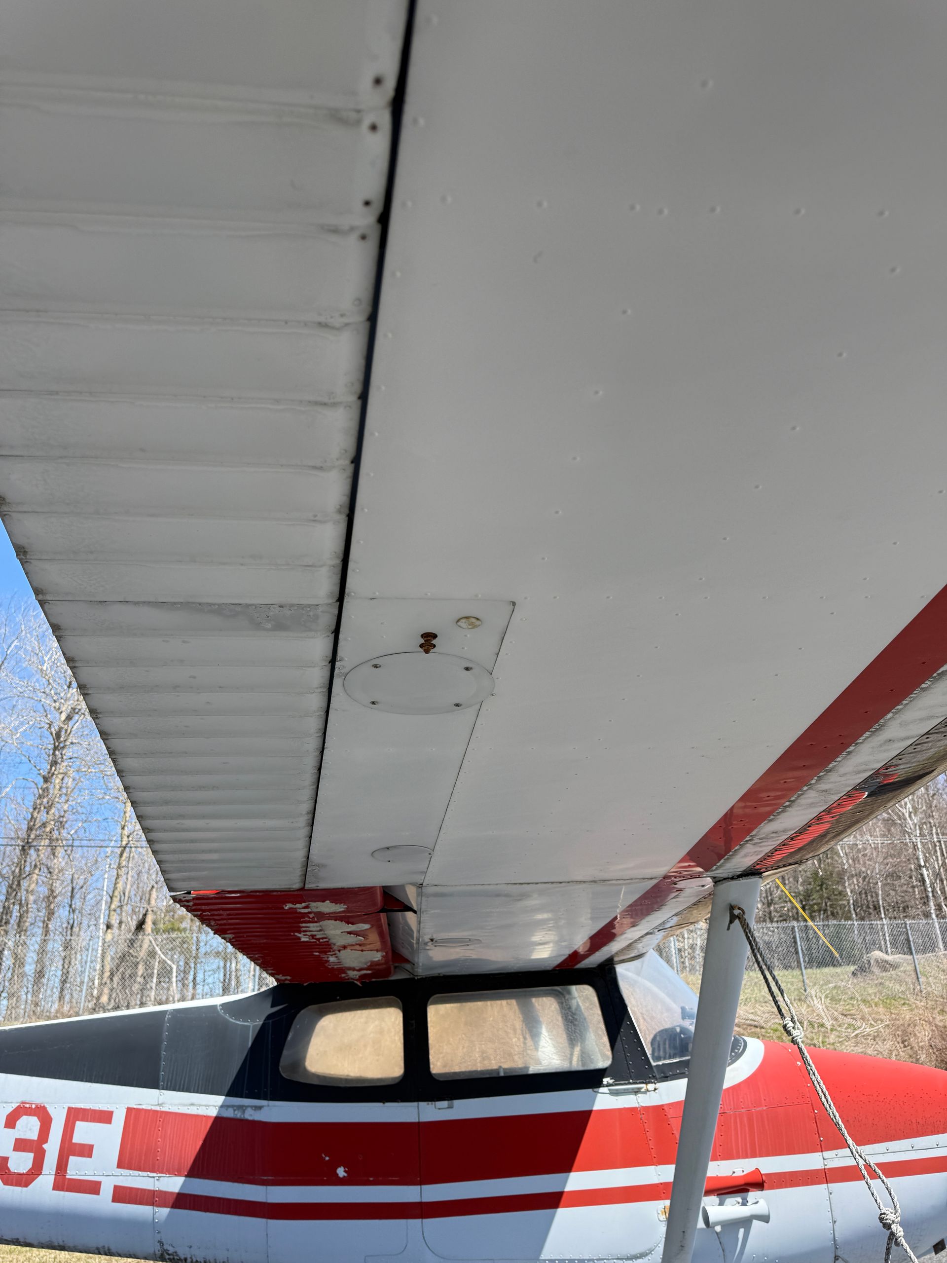 Close-up under a small airplane wing, showing red-and-white fuselage, windows