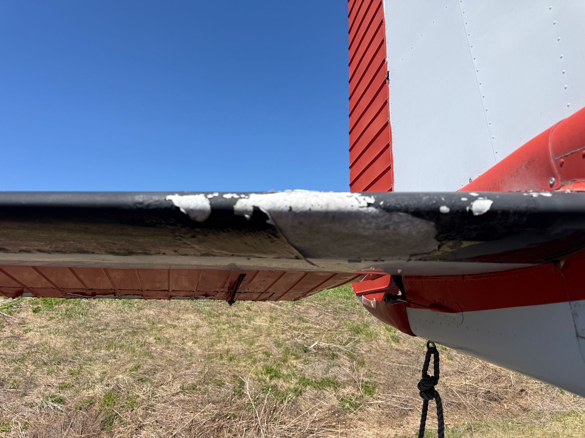 Close-up of an airplane wing with chipped black paint and a red-and-white fuselage against blue sky